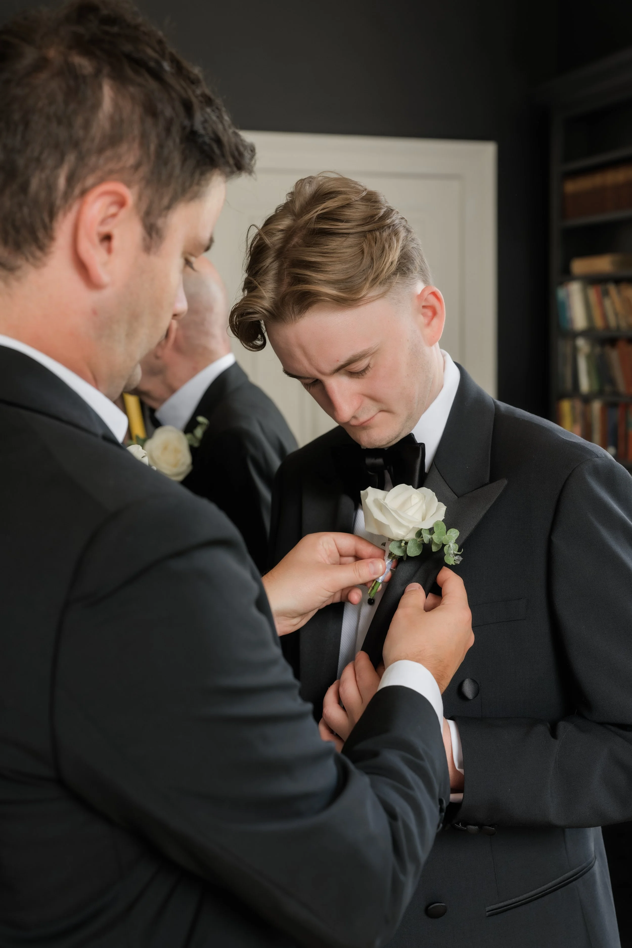 A man in a tuxedo is helping another man in a tuxedo pin a white rose boutonniere on his lapel during a wedding ceremony, with a third man in a tuxedo in the background.