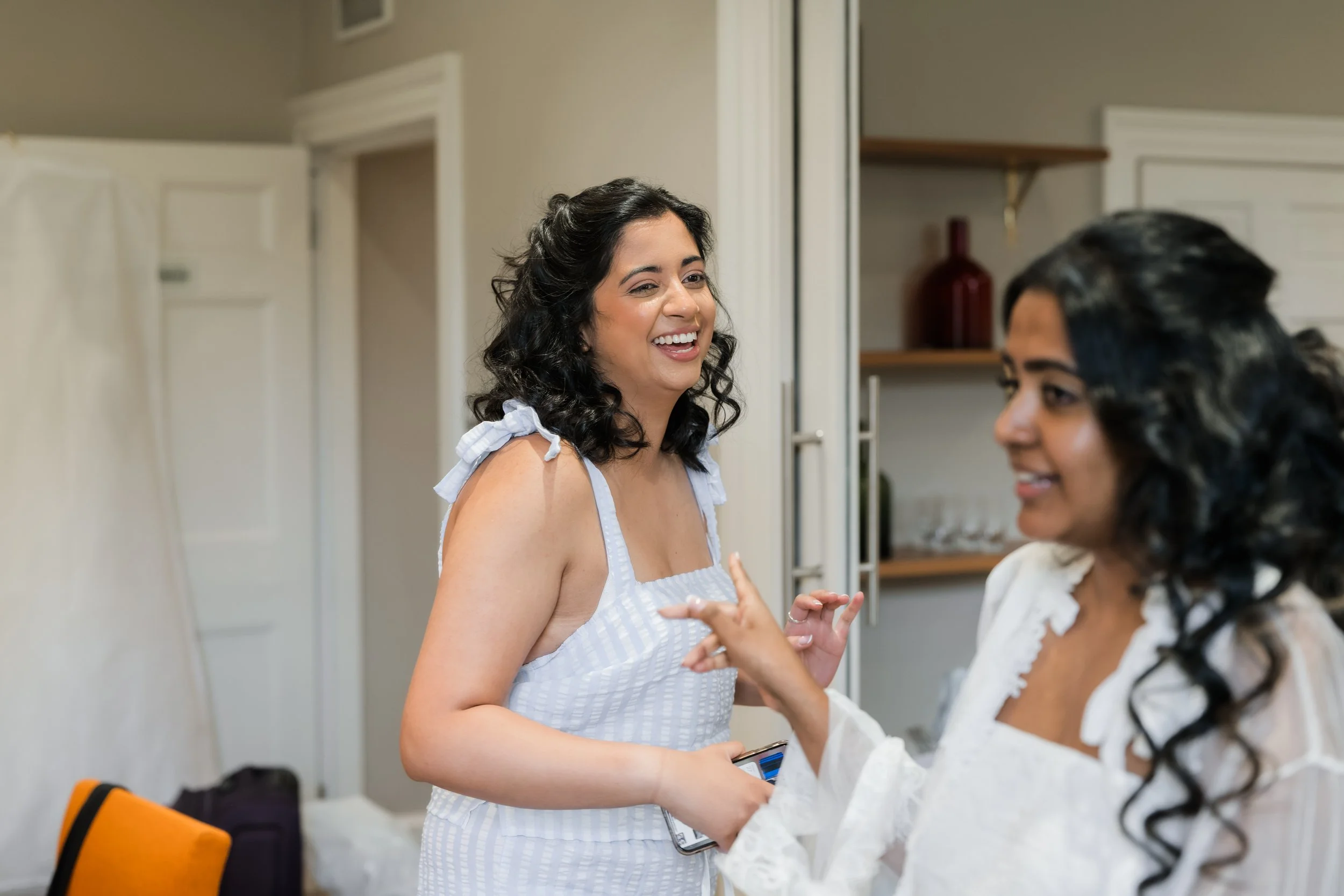 Two women with black curly hair smiling and talking indoors, one holding a phone, wearing white outfits.