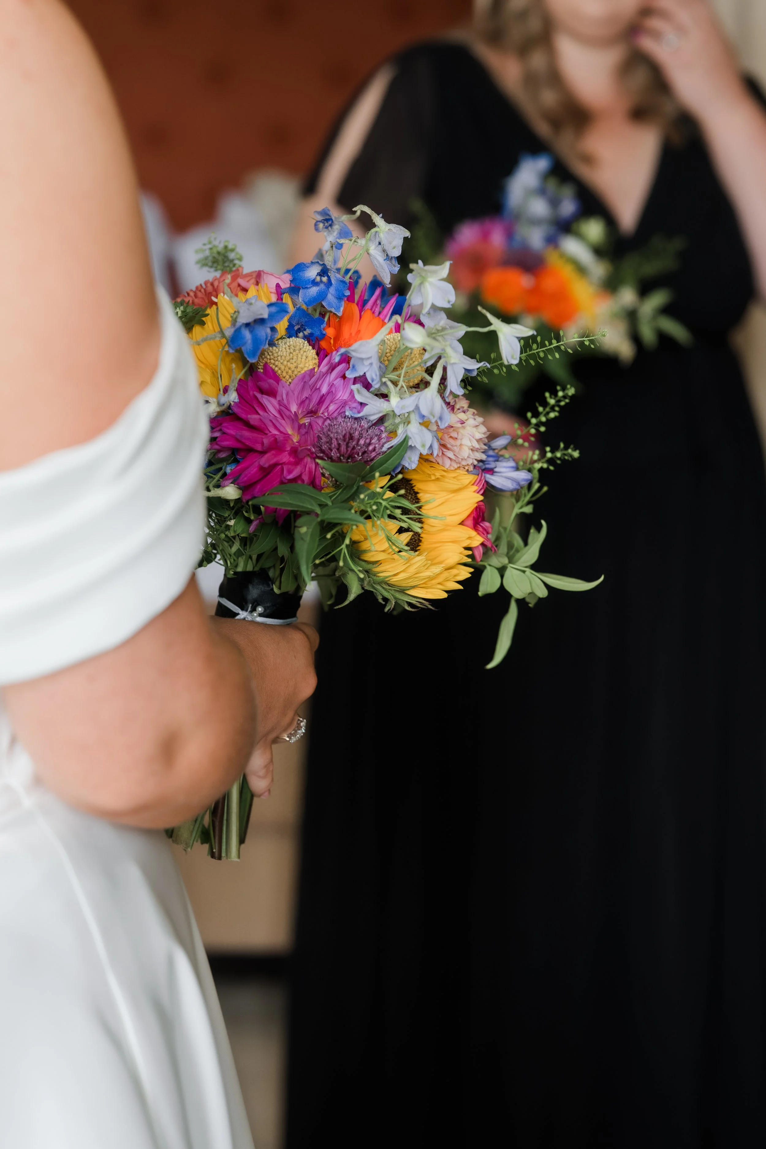 A person in a white dress holding a colorful bouquet of flowers with pink, yellow, and blue blooms, with another person in a black dress holding a bouquet in the background.