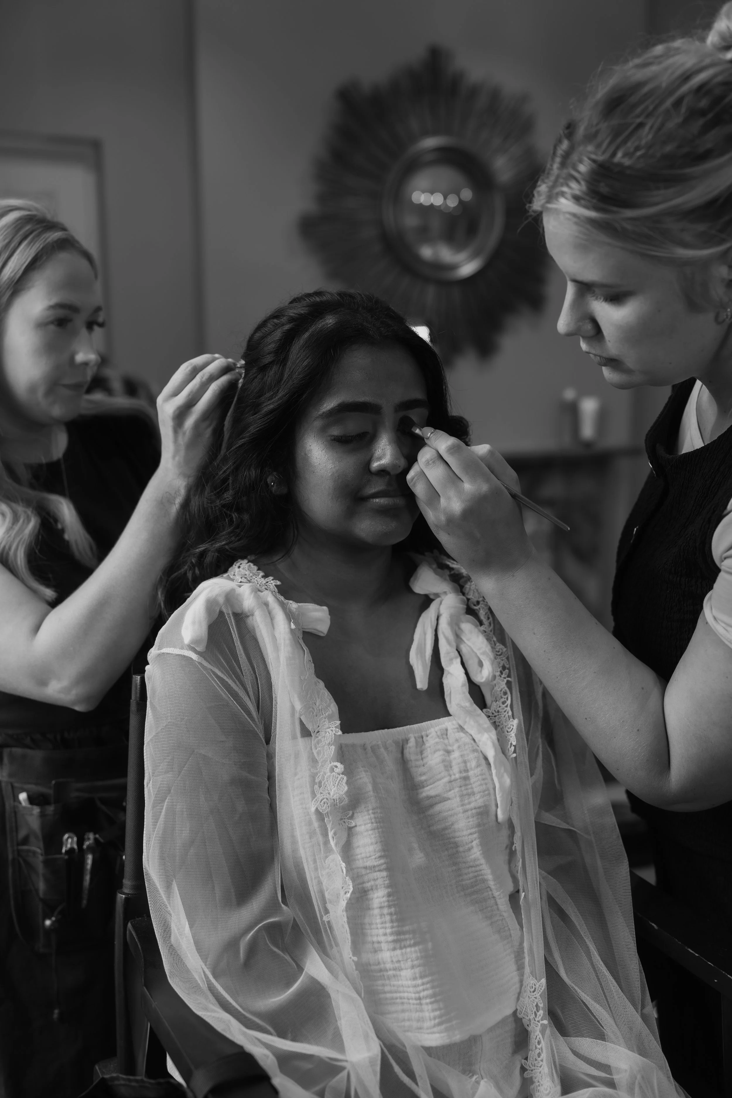 A woman is getting her makeup applied by a makeup artist, with another woman assisting in the background, in a room with a decorative circular mirror on the wall.