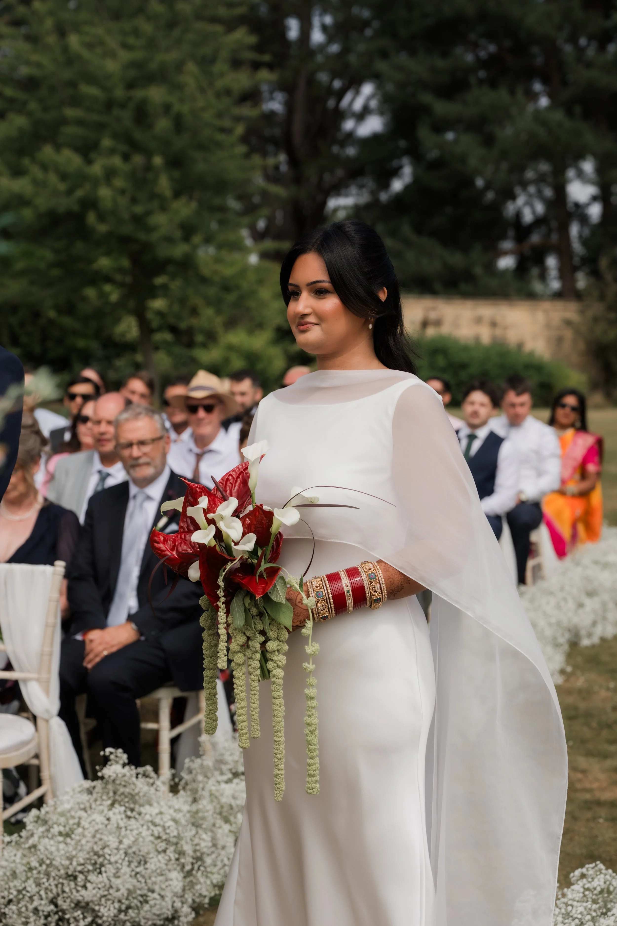 A bride in a white sari holding a bouquet of white and red flowers, with wedding jewelry and bangles, during an outdoor wedding ceremony with seated guests and green trees in the background.