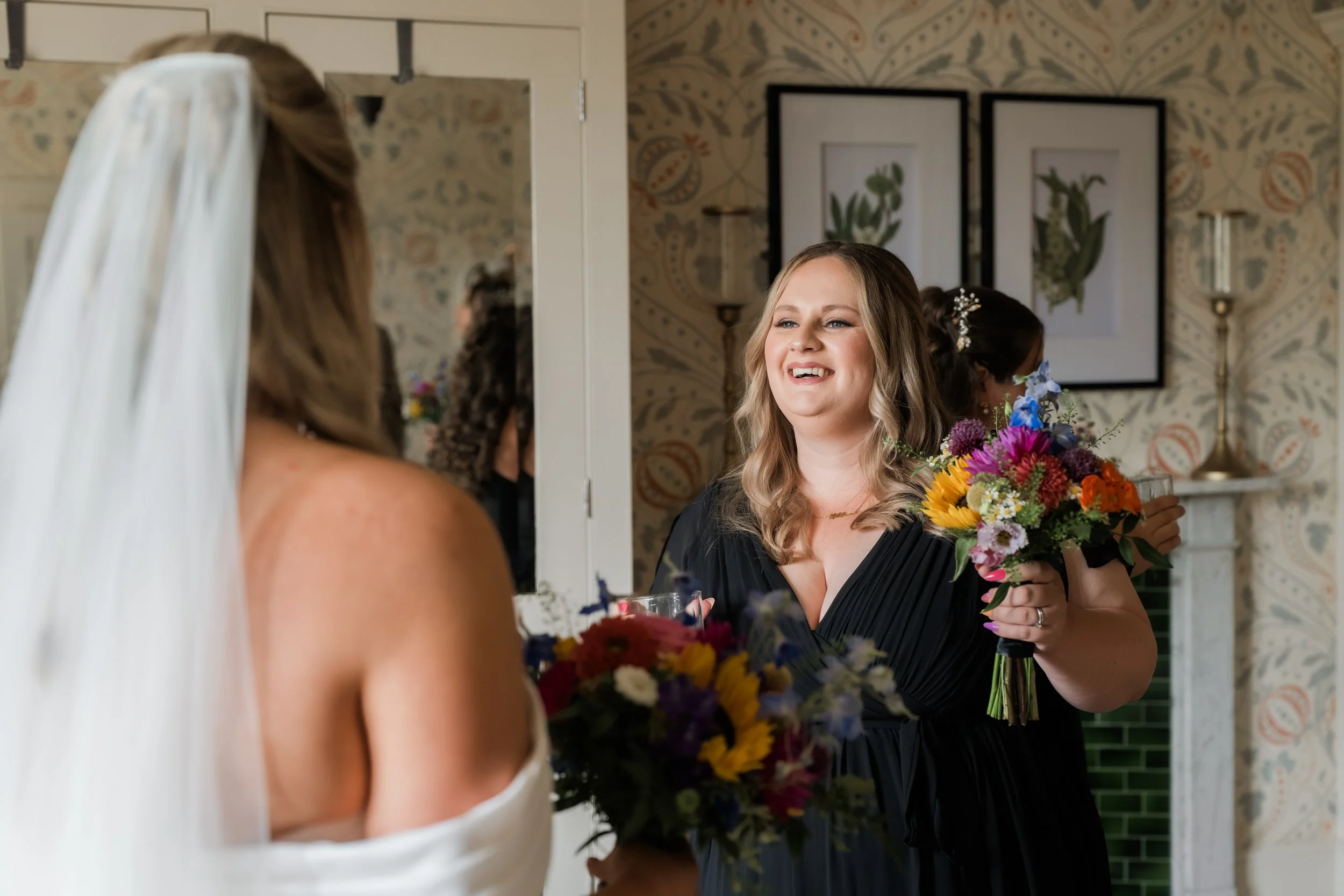 A woman in a black dress holding a colorful bouquet of flowers, smiling and looking at a bride, who is partially visible with a white wedding dress and veil, in a decorated room with framed artwork and a fireplace.