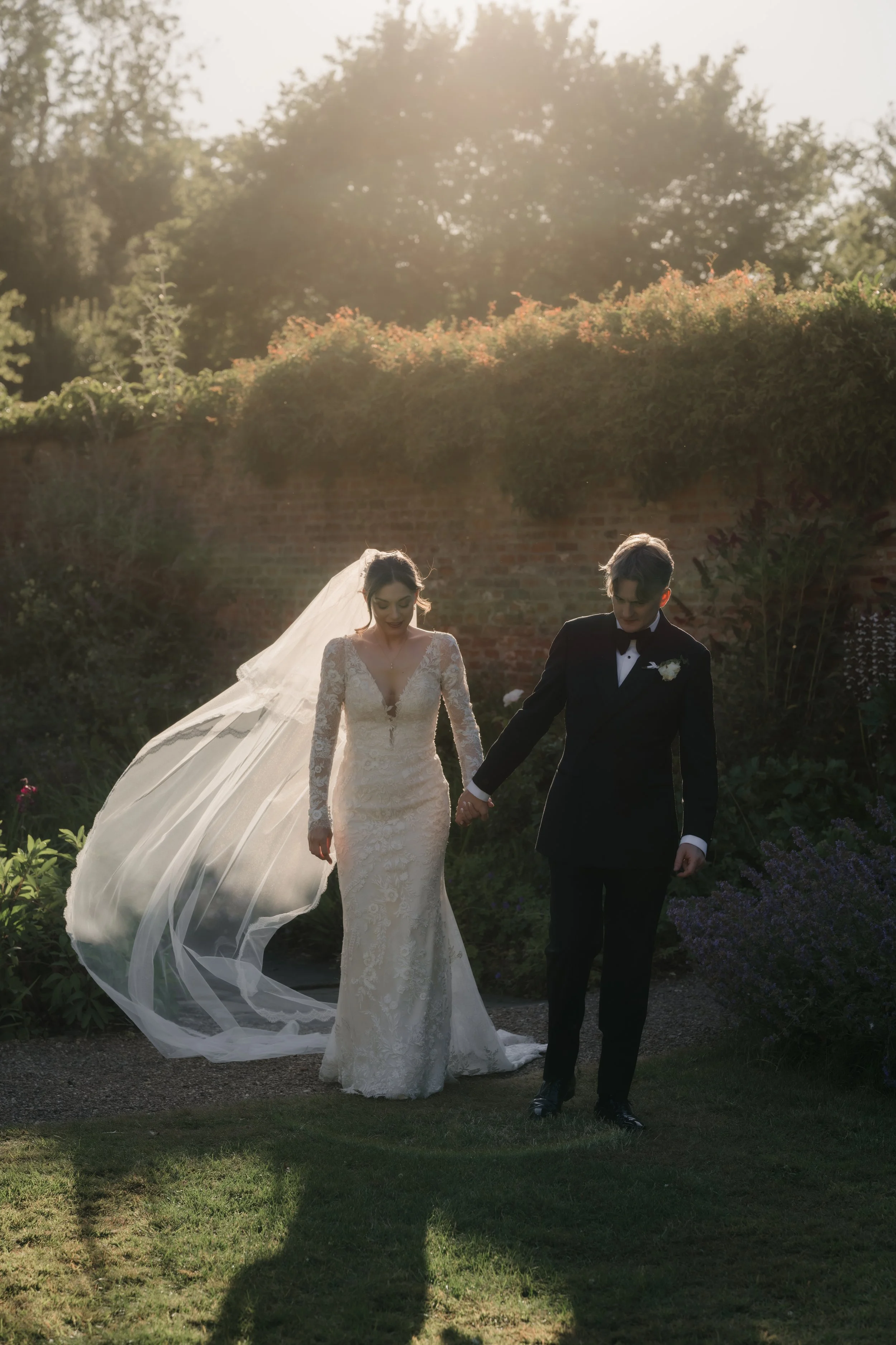 A bride and groom walking outdoors in a garden, holding hands. The bride is in a lace wedding gown with a long veil, and the groom is in a black tuxedo. The scene is illuminated by sunlight, creating shadows on the grass.