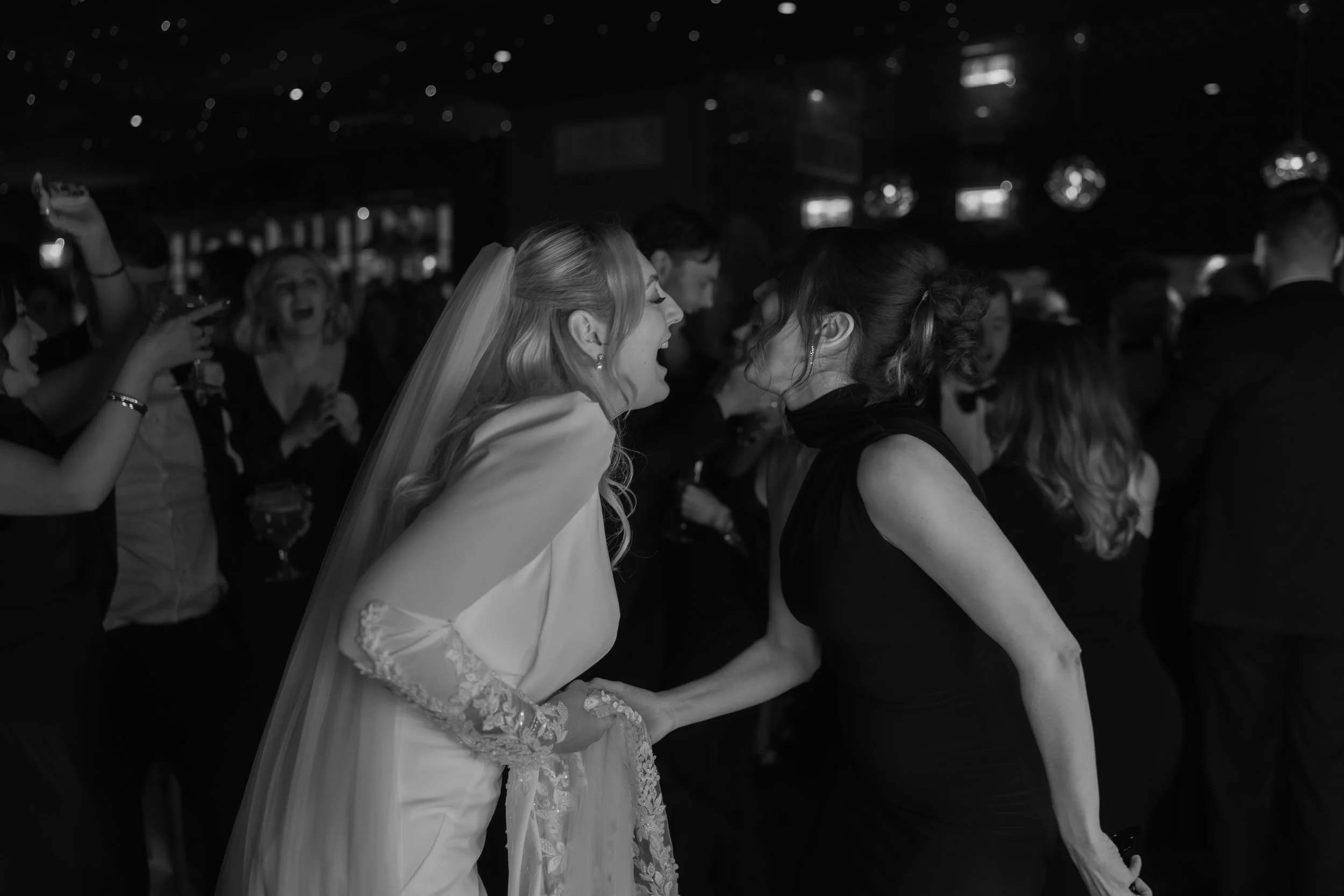 A bride in a wedding dress laughs and leans in to talk to a woman in a black dress at a wedding reception, with other guests visible in the background.