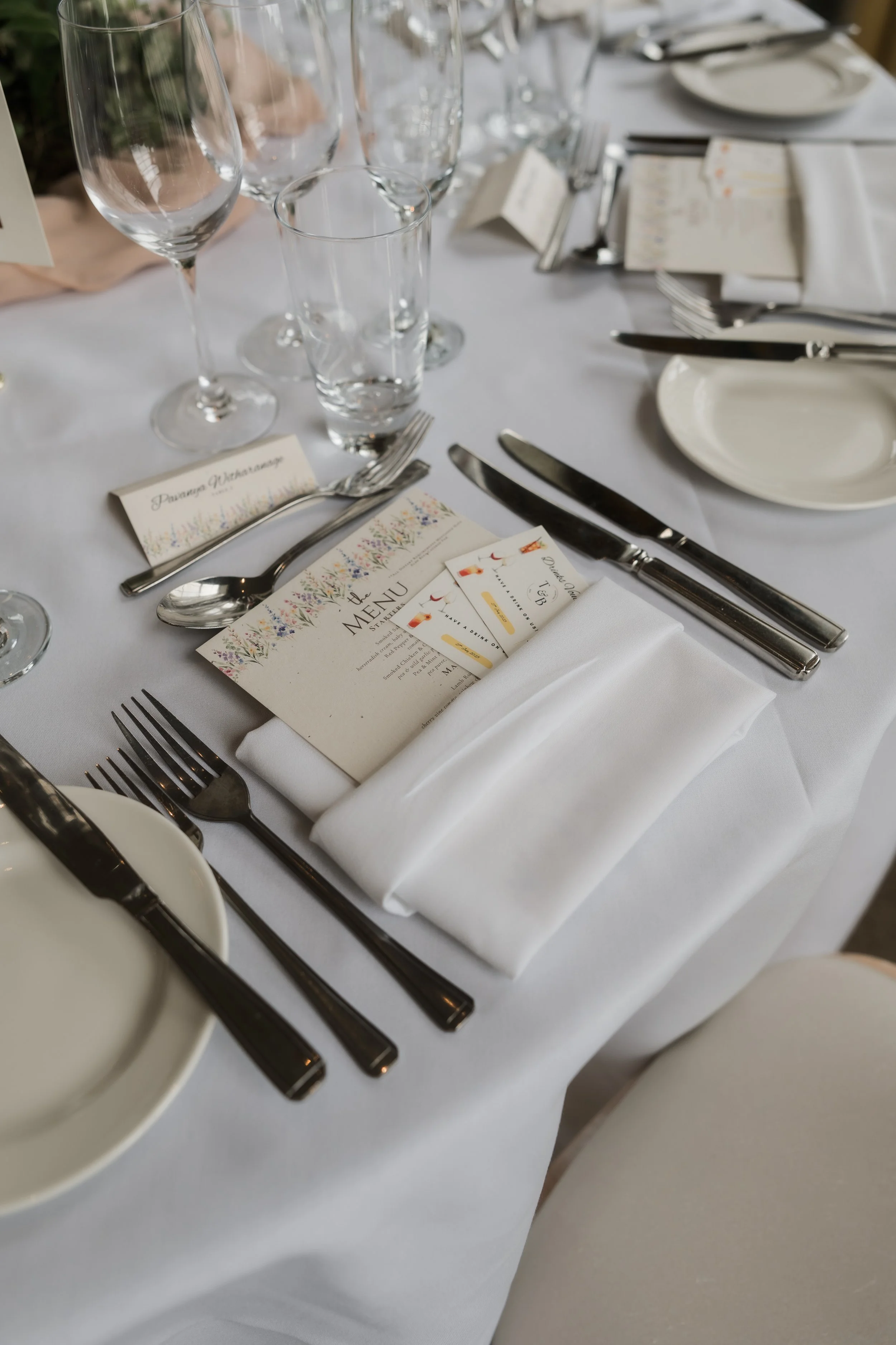 Formal table setting with white tablecloth, multiple glasses, silverware including forks, knives, and spoons, a white napkin, a small plate, and a menu card with colorful floral border.
