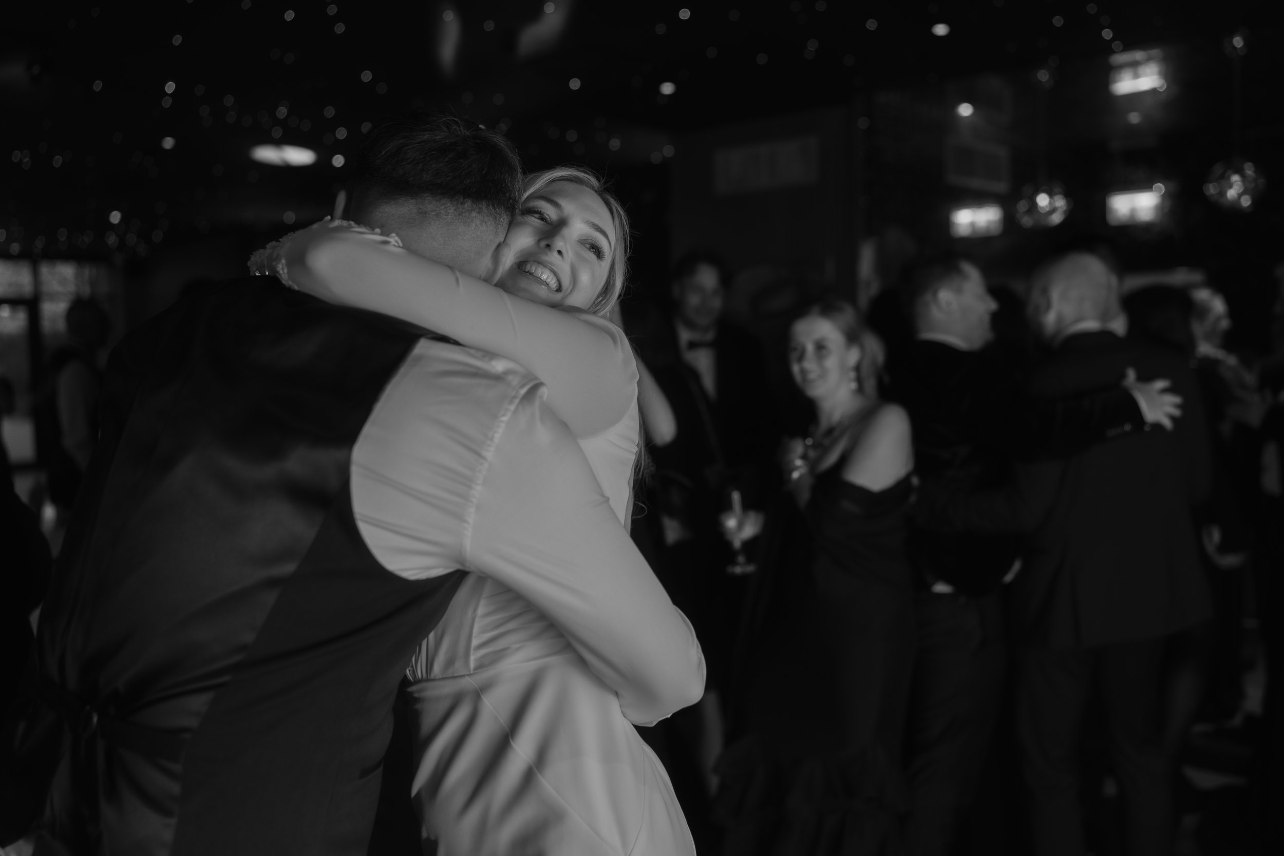 A couple dancing and embracing at a formal event, with other guests in the background holding drinks and talking.