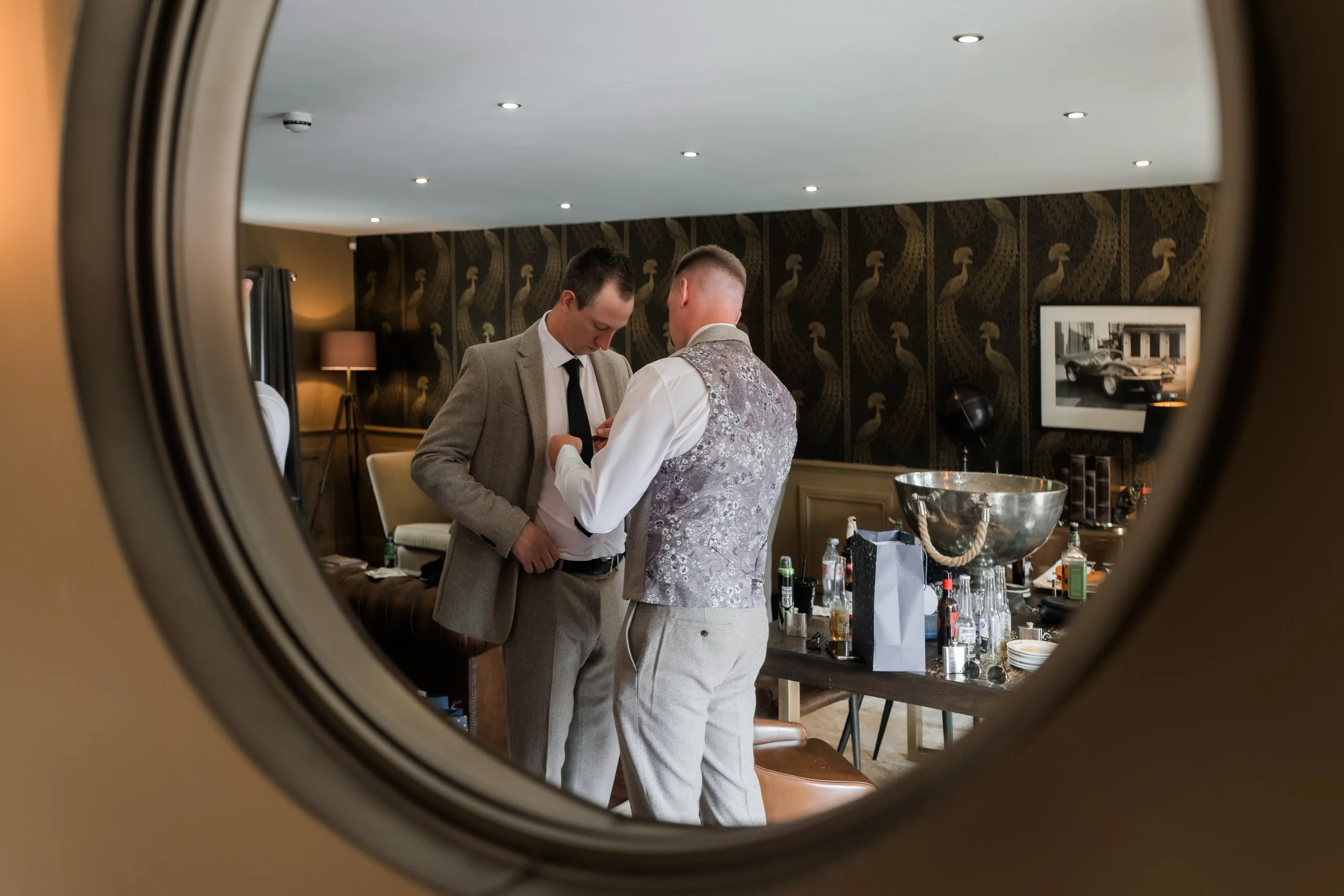 View through a circular mirror showing two men getting dressed in a well-decorated room. One man is adjusting his shirt, the other is helping him. The room has a table with drinks and accessories, and a wall with dark wallpaper and a black-and-white 