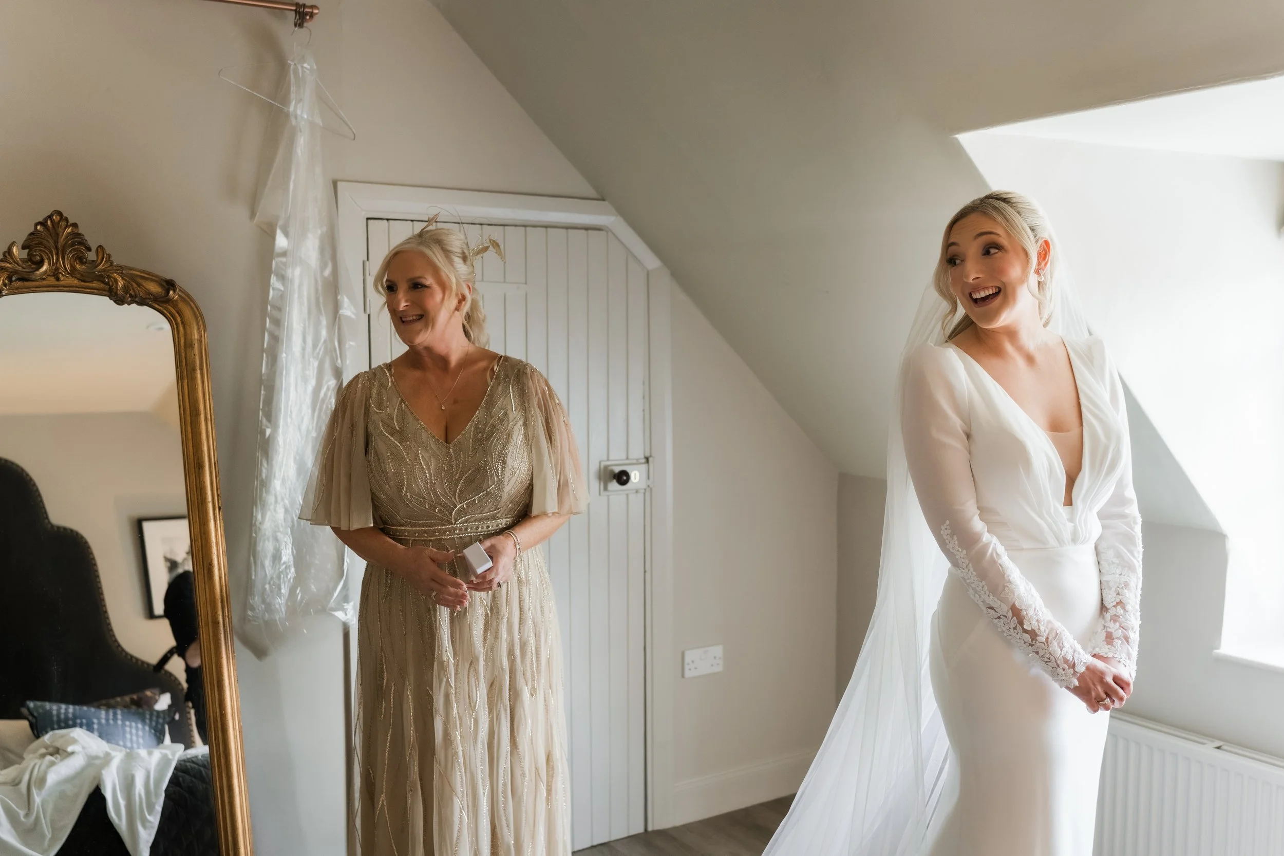 A bride in a white wedding dress with lace sleeves smiling in a bright room next to an older woman in a gold dress, who is looking at her and smiling.