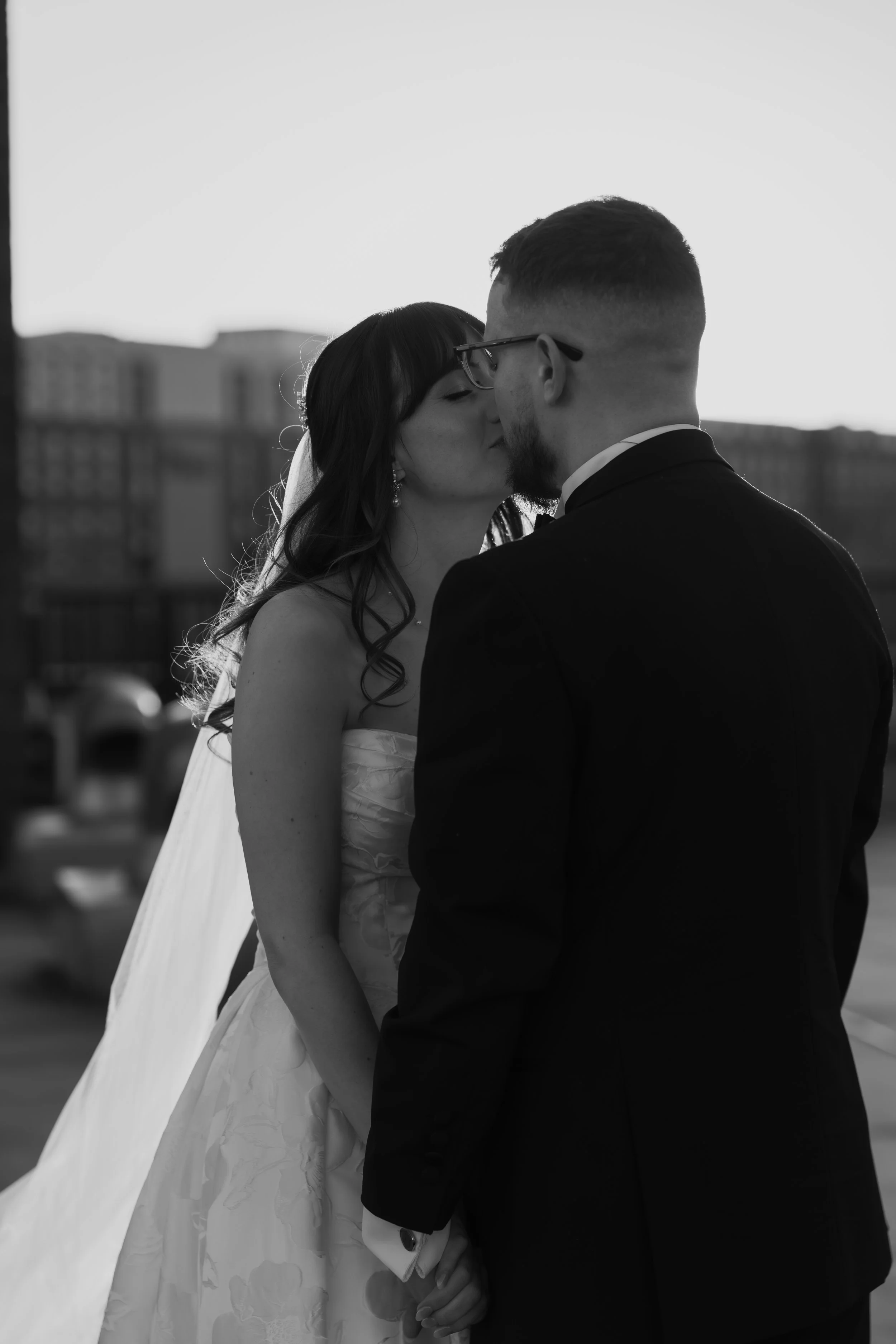A bride and groom sharing a kiss outdoors during their wedding, with city buildings in the background, black and white photo.