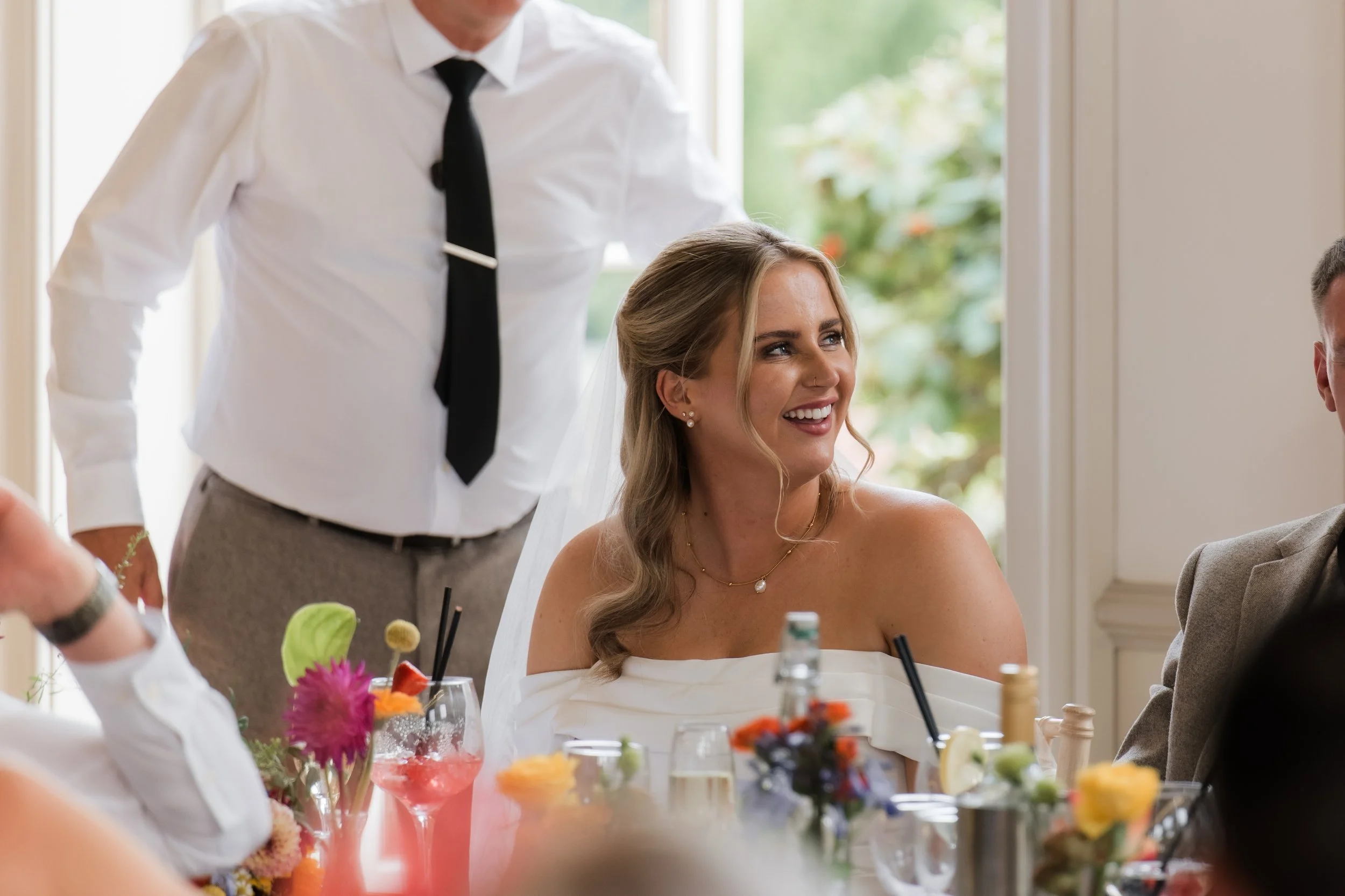 A bride with wavy blonde hair, smiling, wearing a white off-the-shoulder dress and pearl jewelry, sitting at a table during a wedding reception, with a man in a suit seated to her right and a person standing behind her.