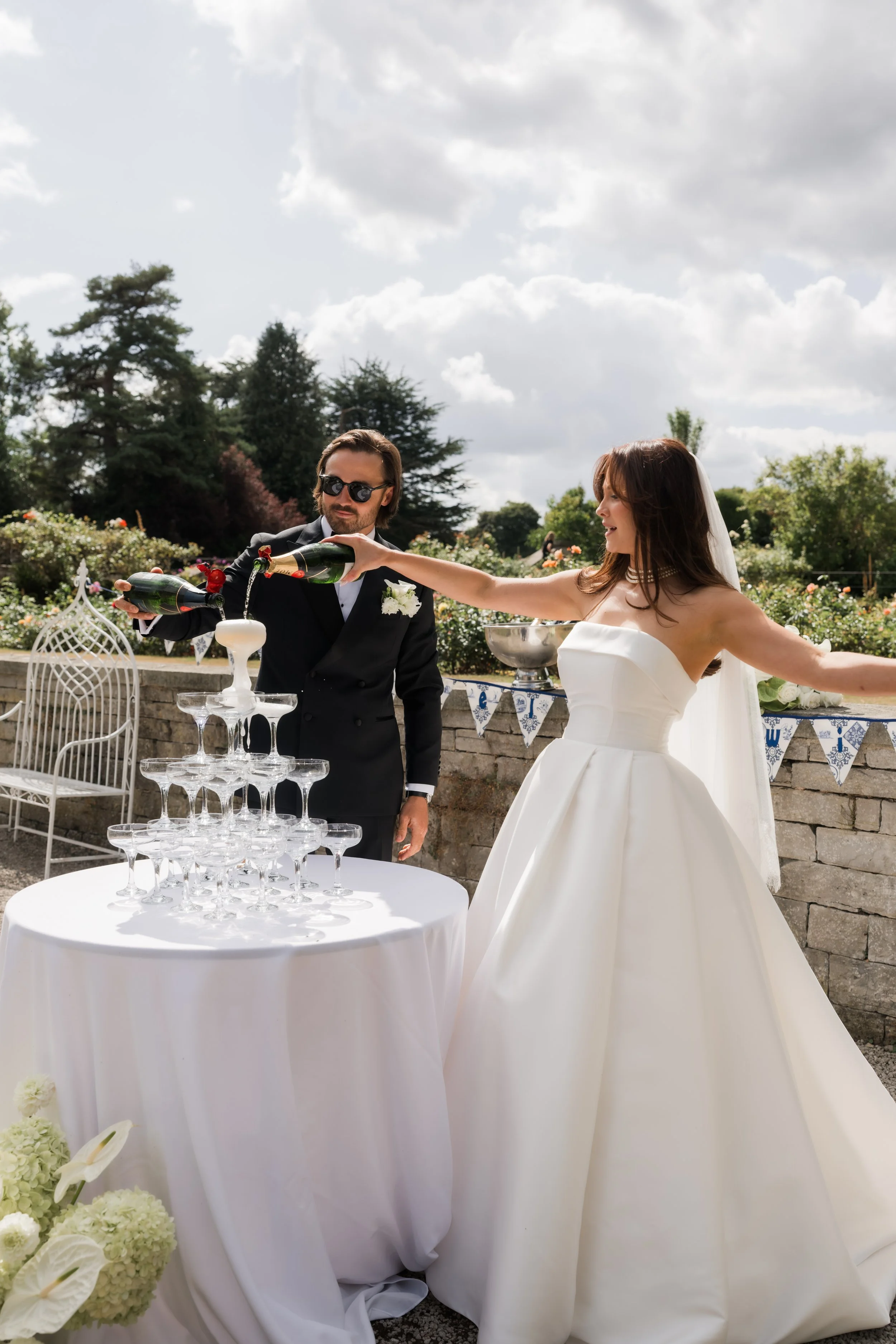 A newly married couple in wedding attire at an outdoor celebration, with the groom pouring champagne into a pyramid of glasses.
