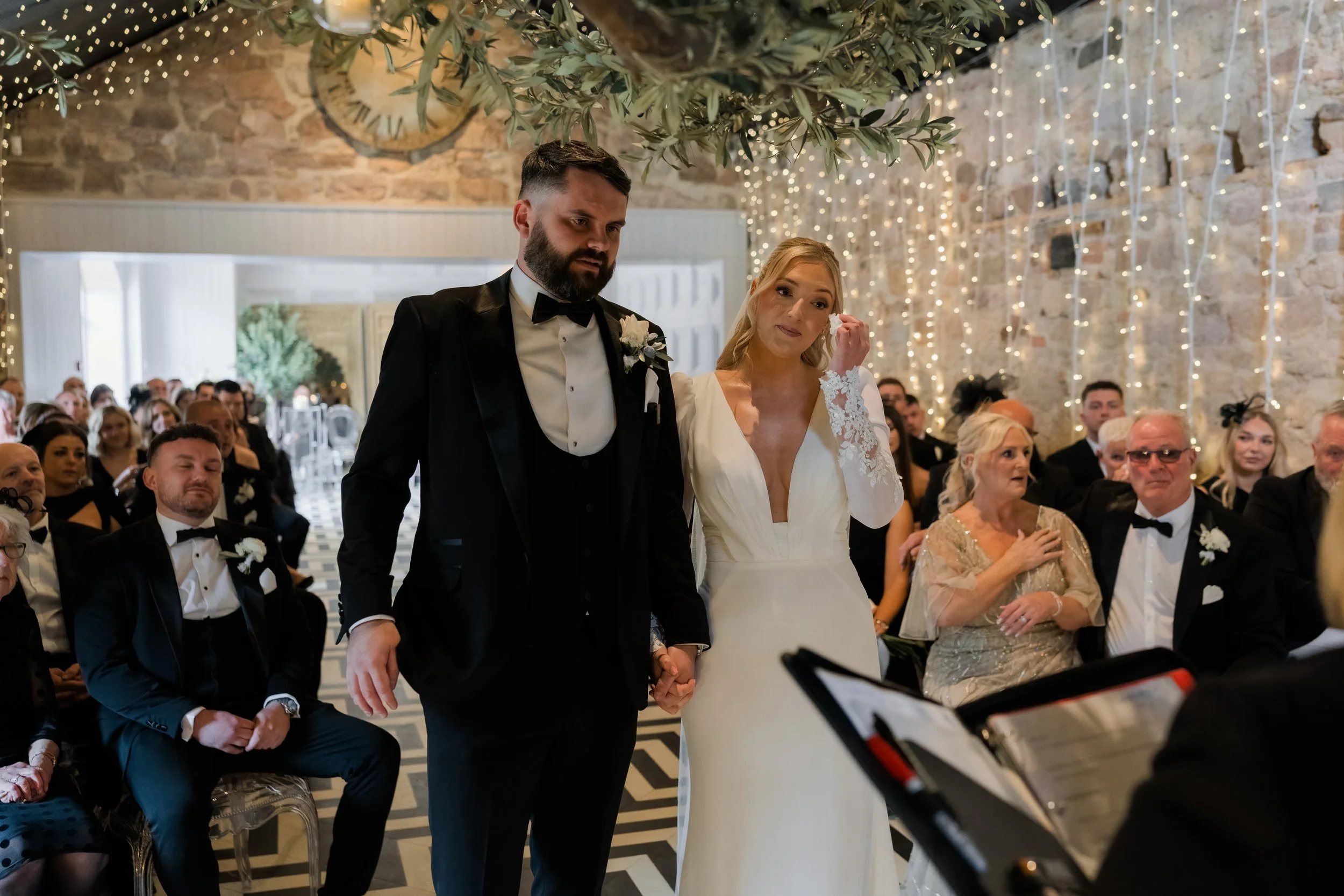 A bride and groom holding hands during their wedding ceremony in a decorated venue, with guests in formal attire watching. The bride is wiping her tears, and the groom looks solemn. The background features fairy lights and a stone wall.