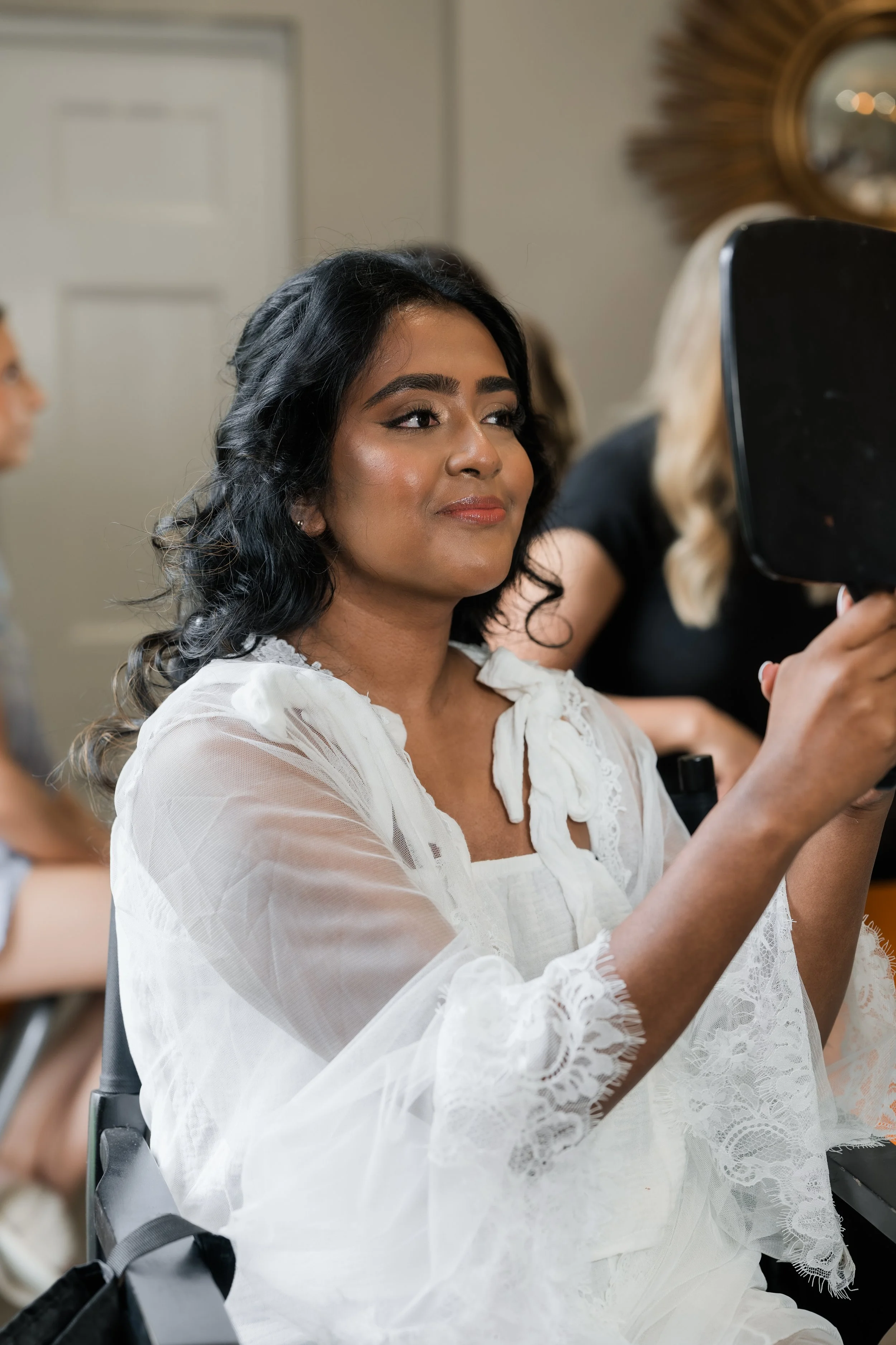 A woman with dark hair and makeup sitting in a makeup chair, looking at herself in a mirror, wearing a white lace blouse.