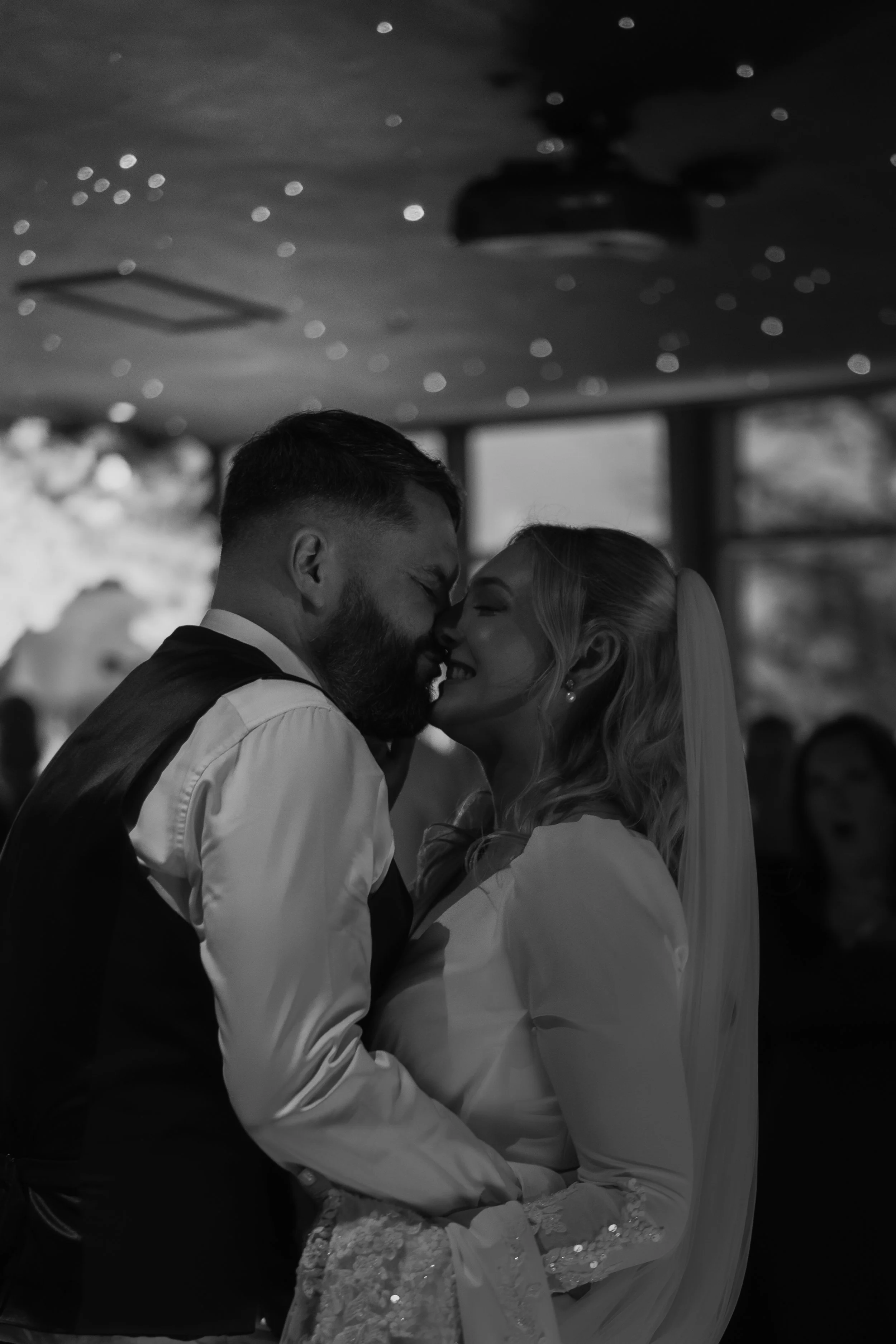Couple sharing a kiss at a wedding reception, with a ceiling decorated with string lights, in black and white.