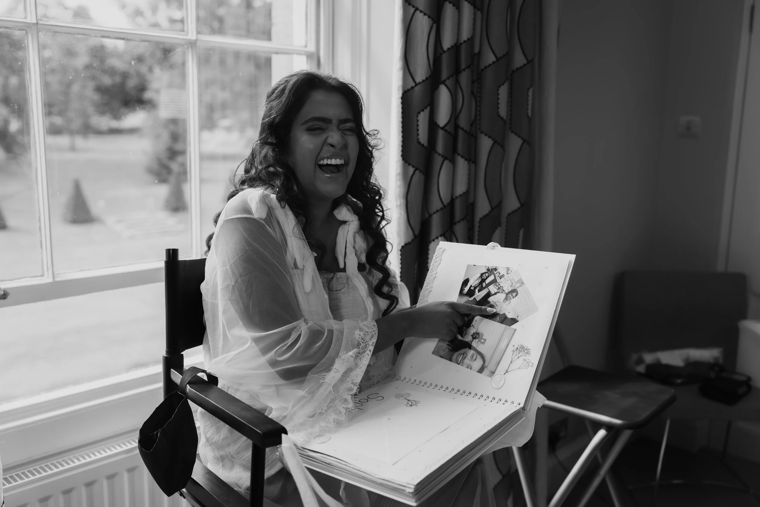 A woman sitting by a window, smiling and pointing at photos in a scrapbook.