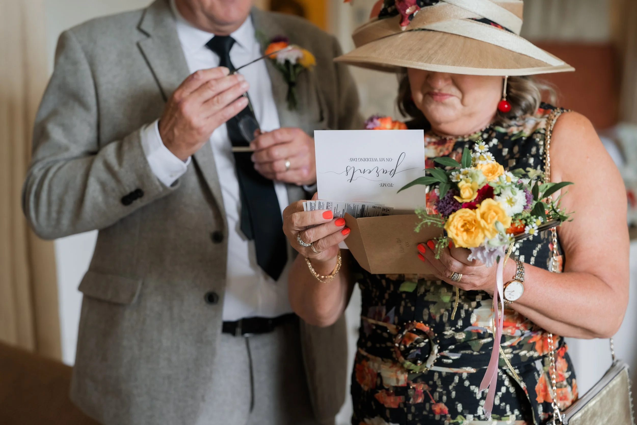 A woman wearing a large sun hat, floral dress, and red lipstick holding a bouquet of flowers and reading a card, while a man in a gray suit with a boutonniere stands nearby at a wedding celebration.