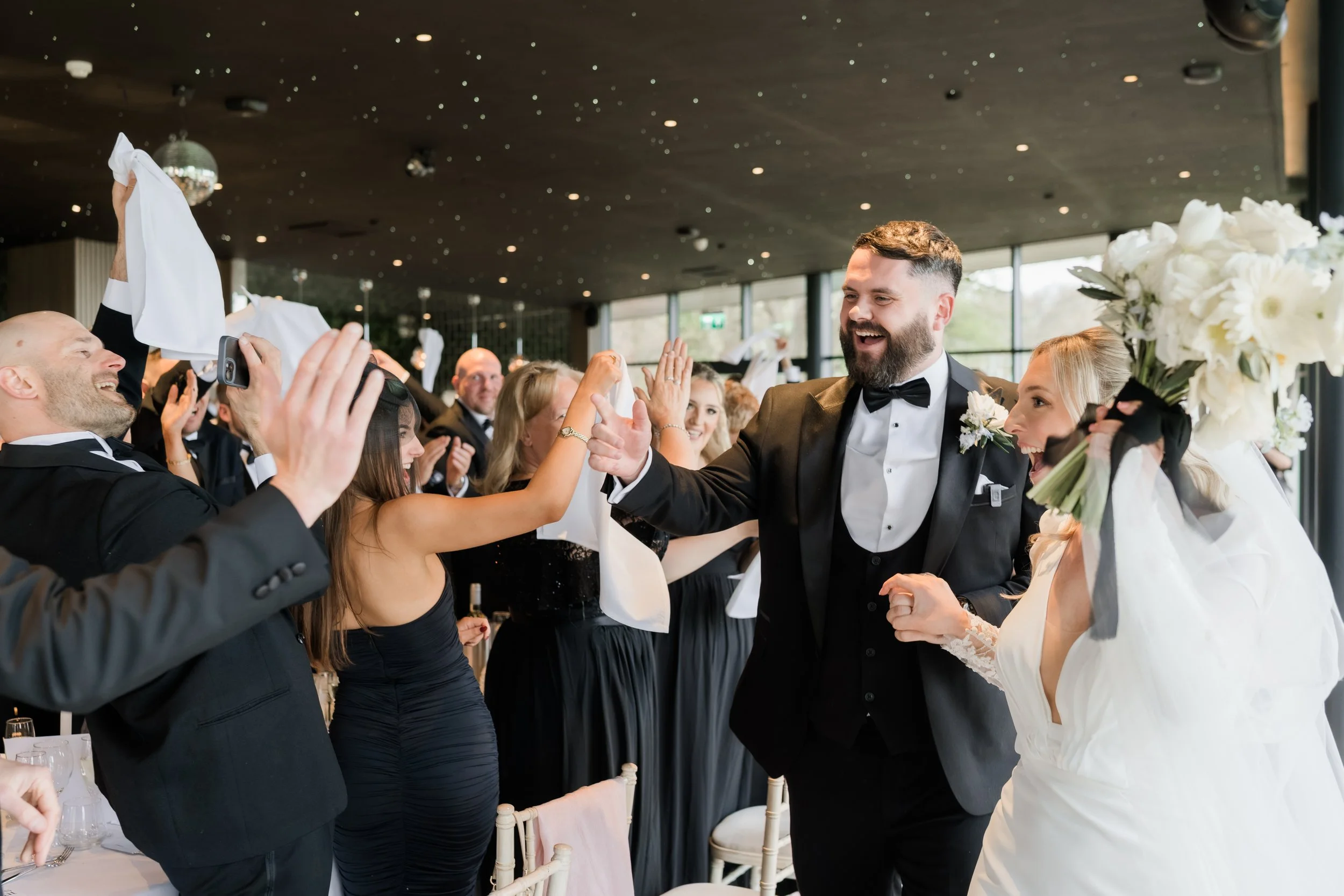 Wedding reception with bride and groom high-fiving, guests taking photos, indoors with large windows and ceiling lights.