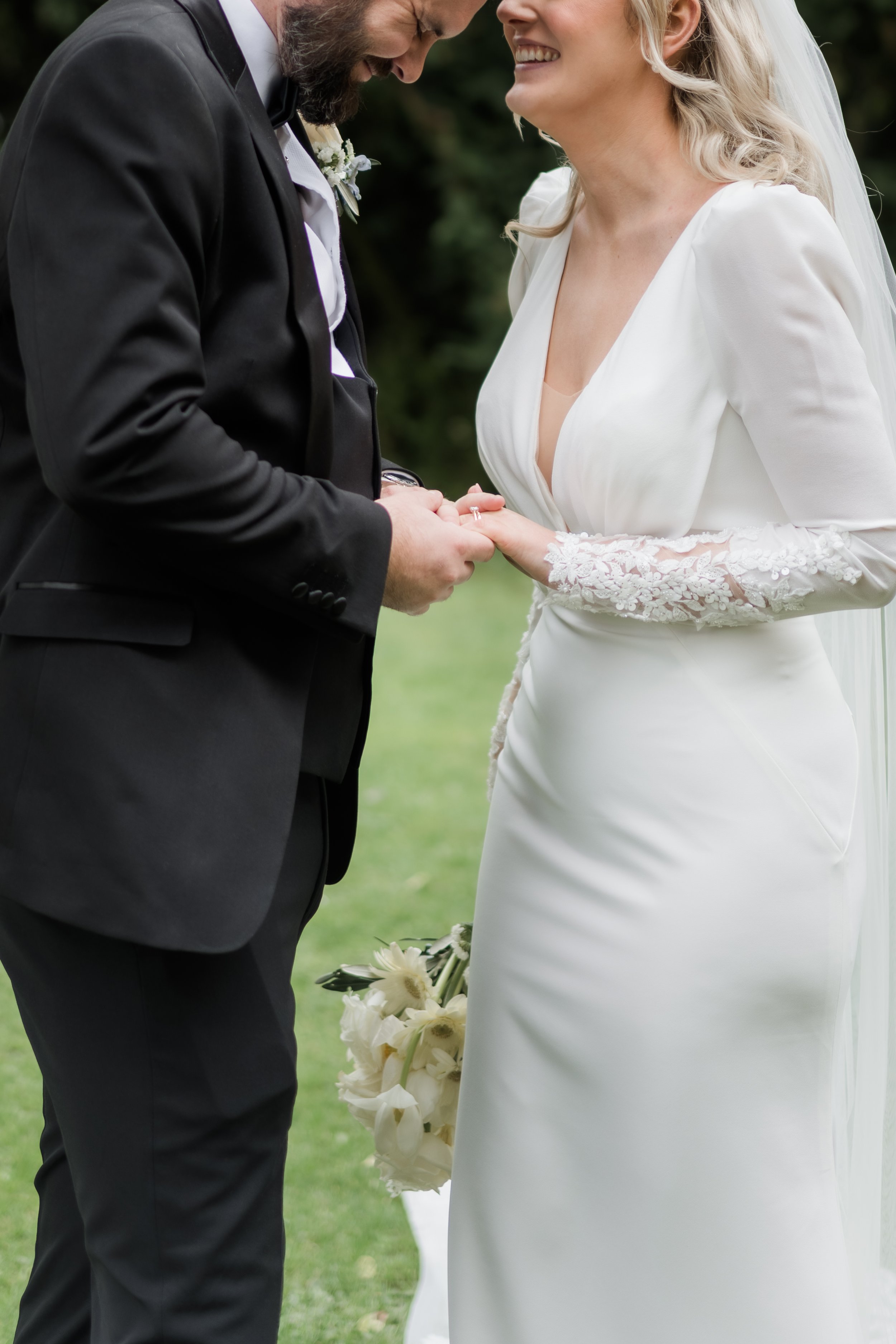 A bride and groom exchanging wedding rings outdoors, with the bride holding a bouquet of white flowers, both smiling and looking at each other.