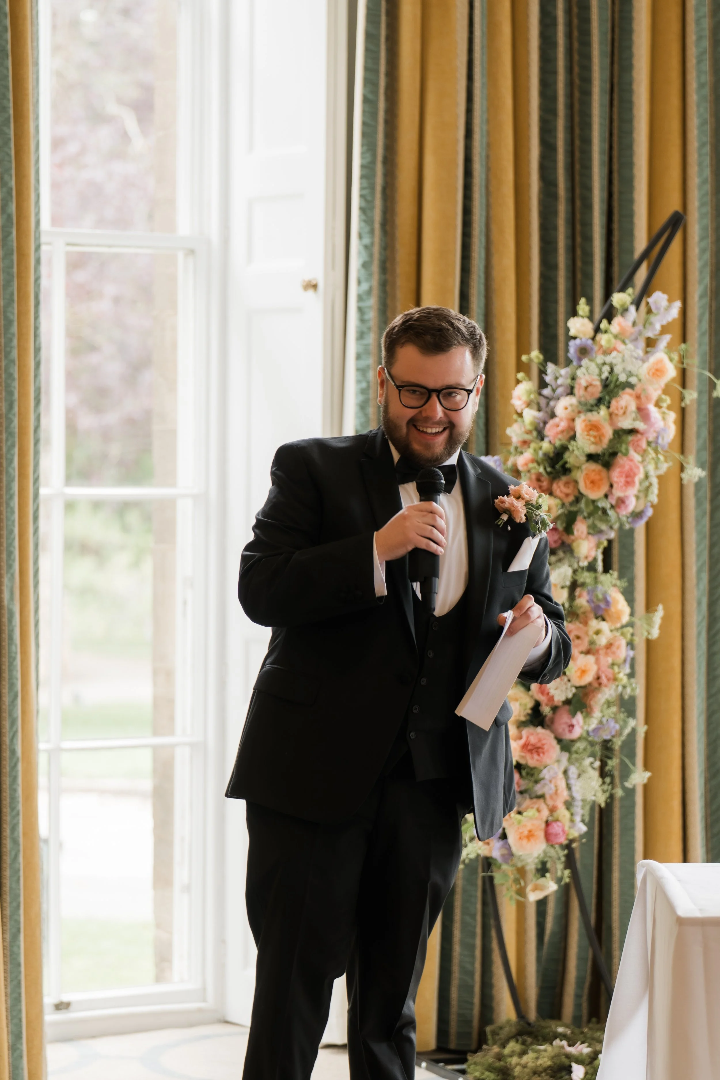 A man in a tuxedo speaking into a microphone during a wedding or formal event, standing in front of a large window and floral arrangement.