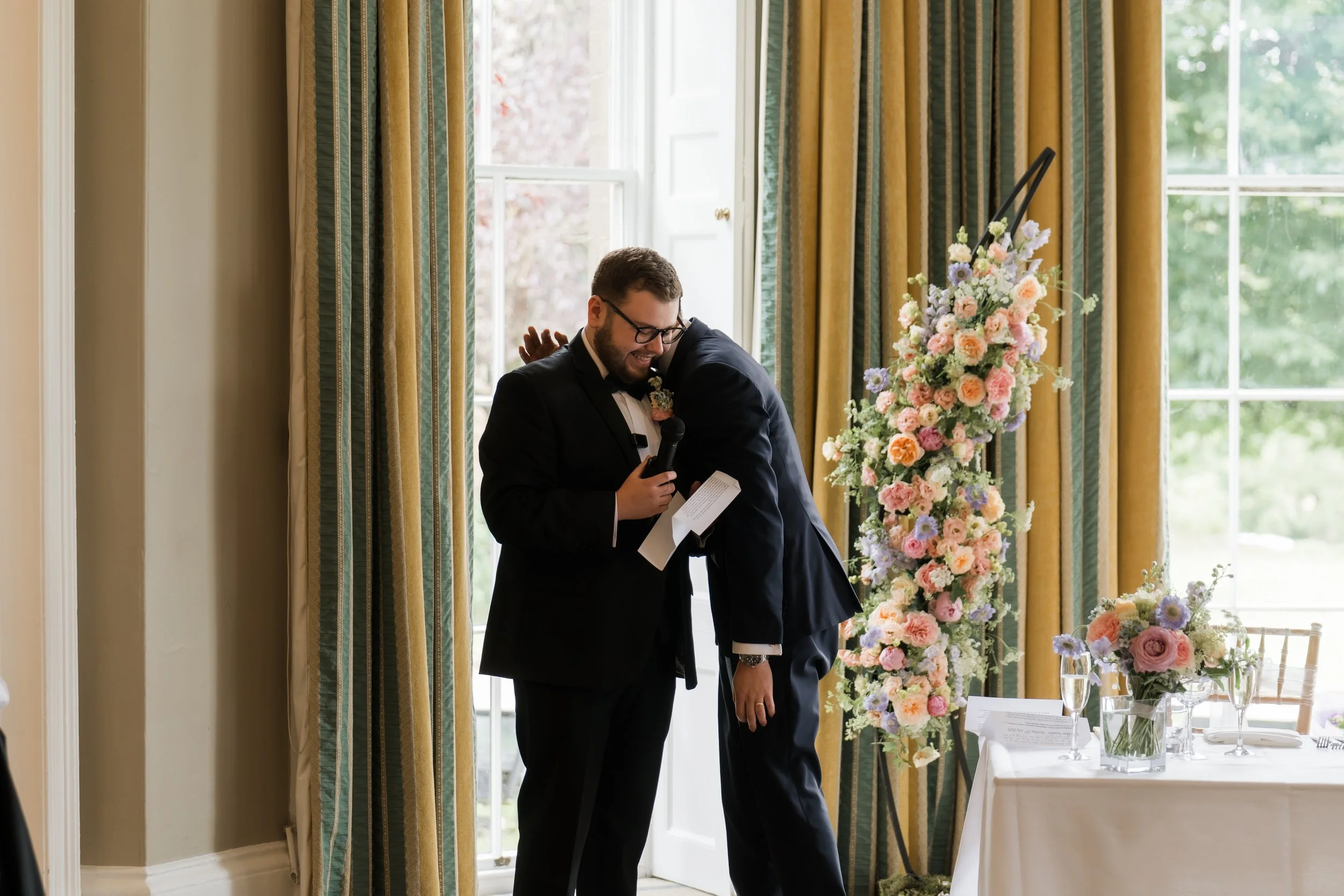 Two men dressed in black suits, one with a beard and glasses, hugging at a wedding reception near a window with yellow curtains and pink and purple floral arrangements on a table.