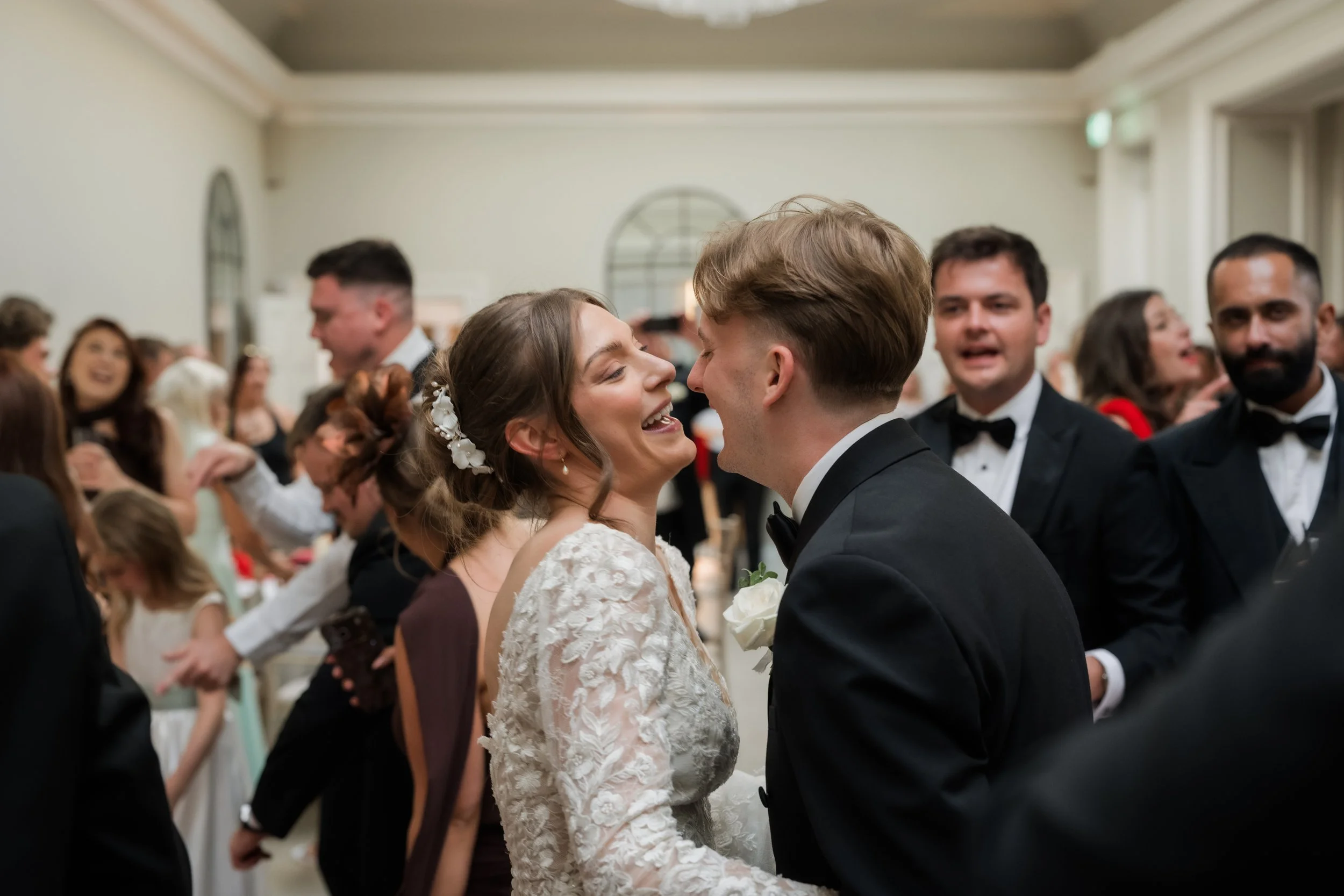 A bride and groom are smiling and holding hands at their wedding reception, surrounded by guests.