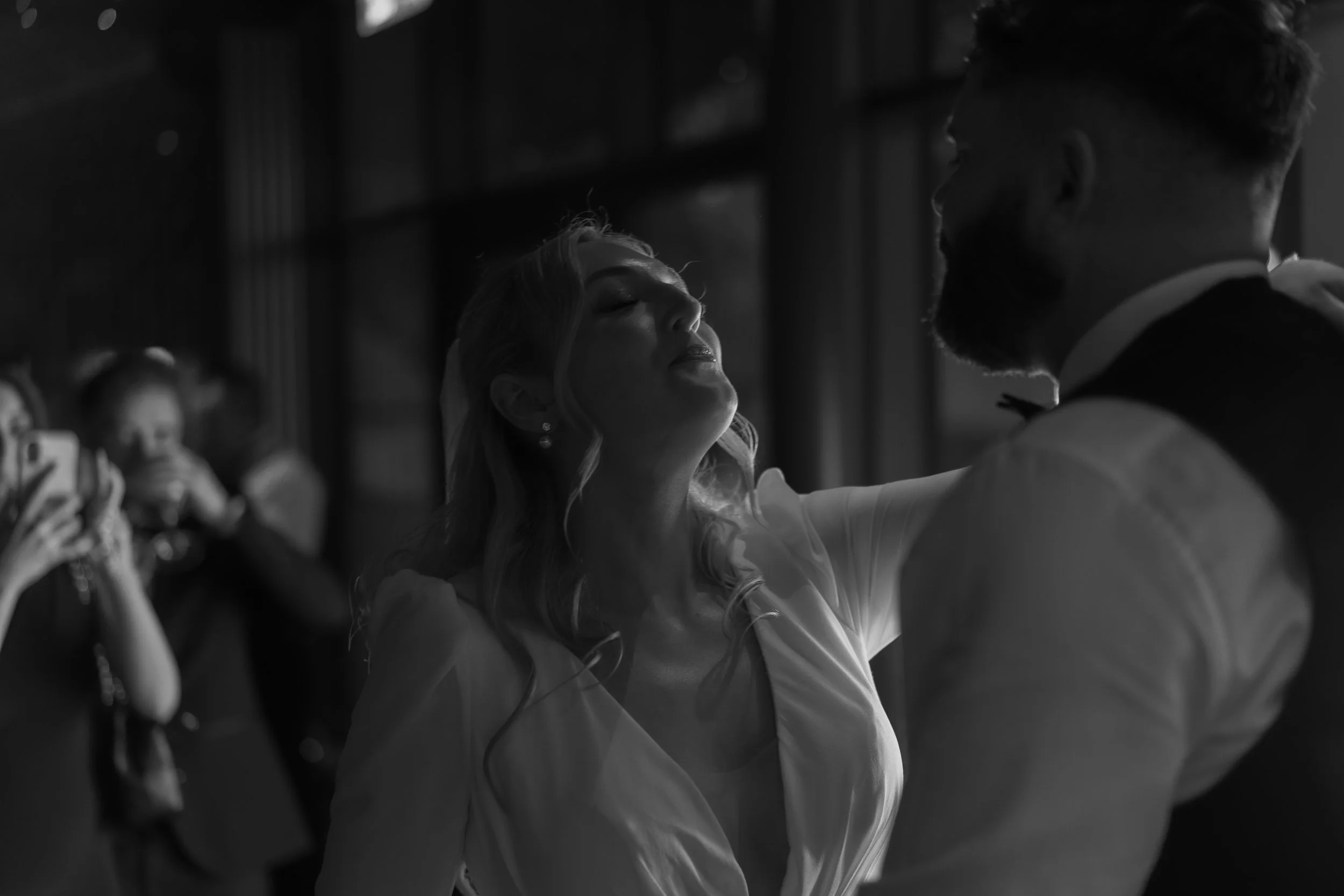 A woman and a man dancing closely at a social event, with women in the background taking photos.