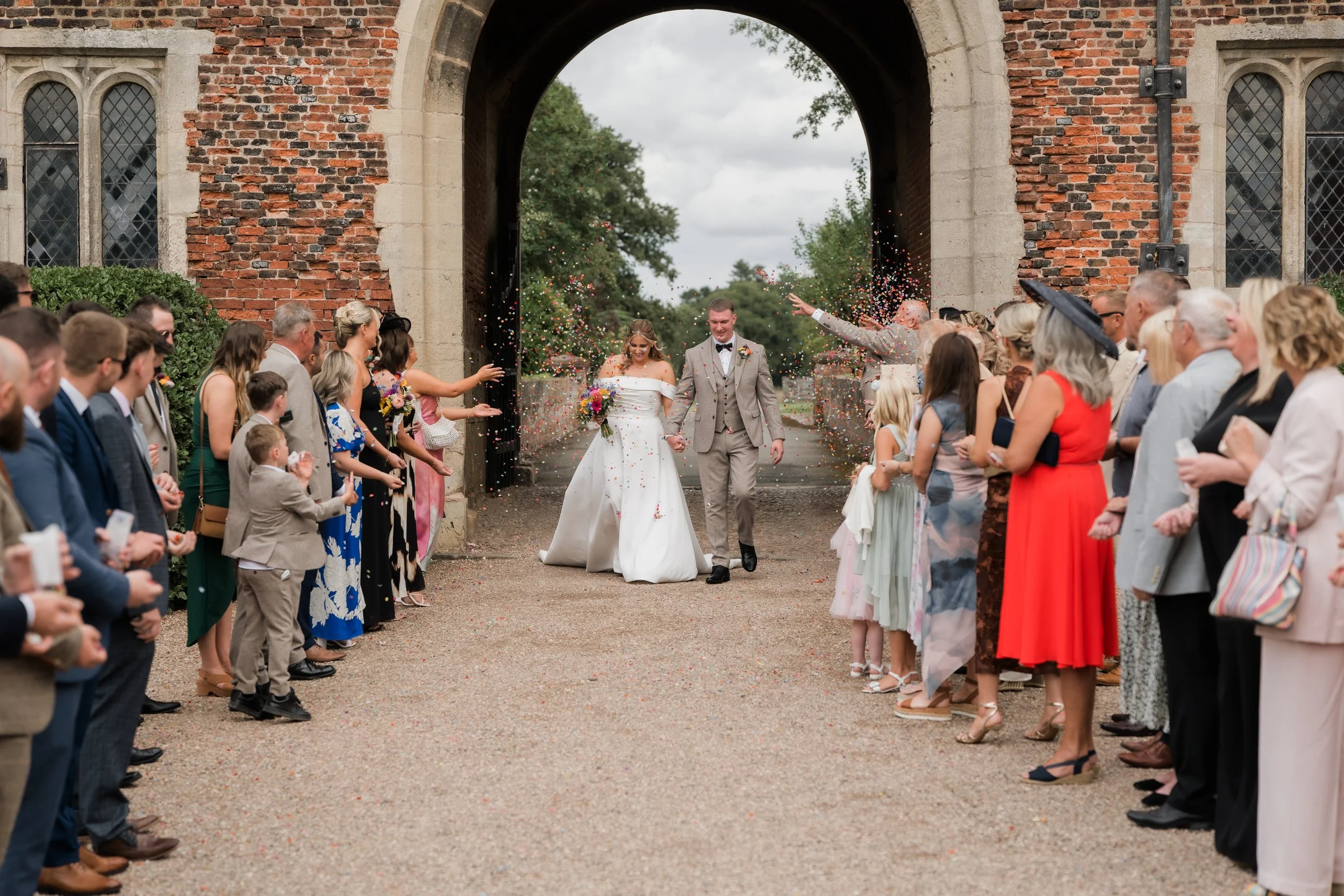 A newlywed couple walking hand in hand through a crowd of wedding guests as they exit a brick archway, with confetti falling around them.