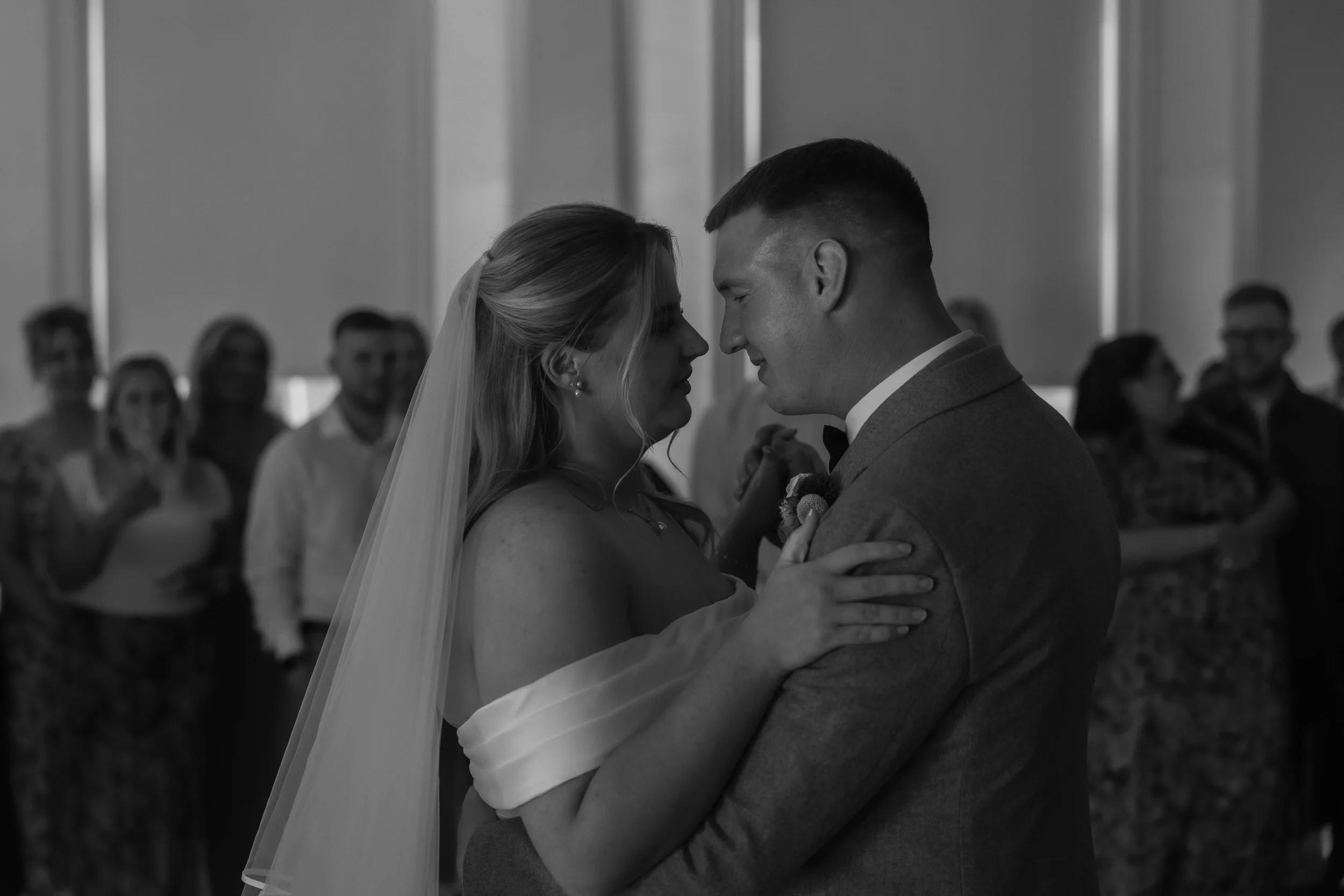 A bride and groom share a close moment during their wedding dance, with wedding guests blurred in the background.