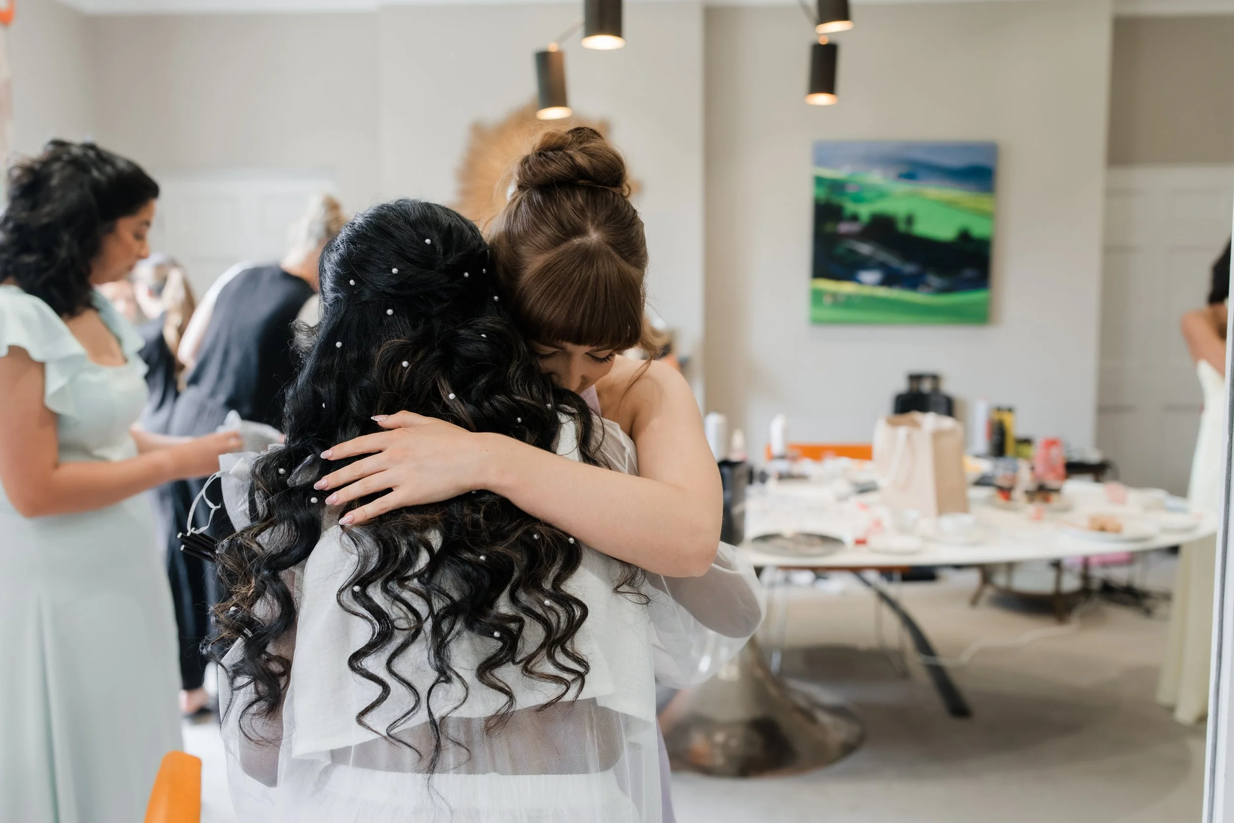 Two women embrace warmly in a room with wedding preparations, other women and a cluttered table in the background.