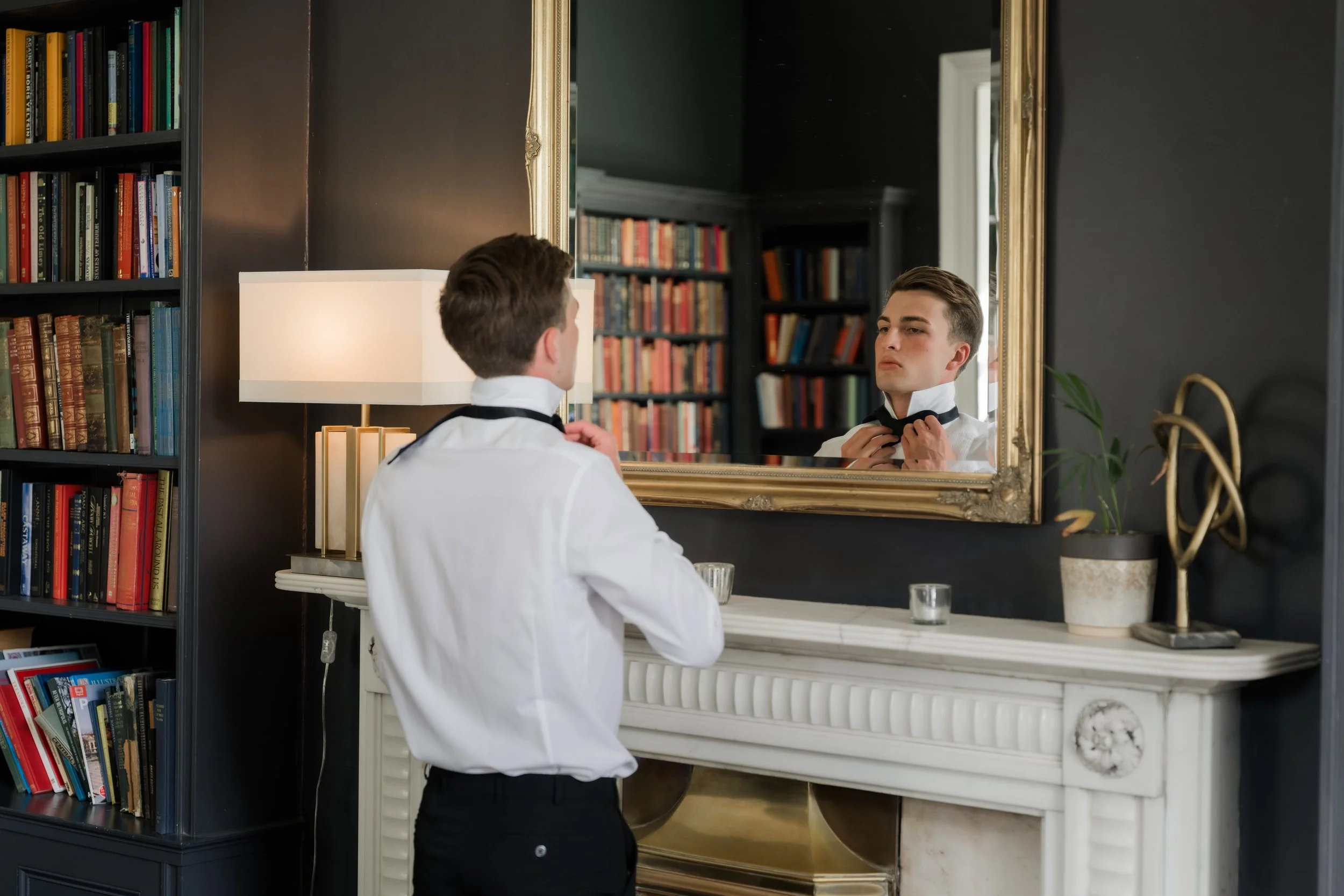 A young man dressed in a white shirt and black trousers is tying a black bow tie in front of a large mirror decorated with a gold frame. The room has dark walls, a tall bookshelf filled with colorful books on the left, a white marble fireplace on the