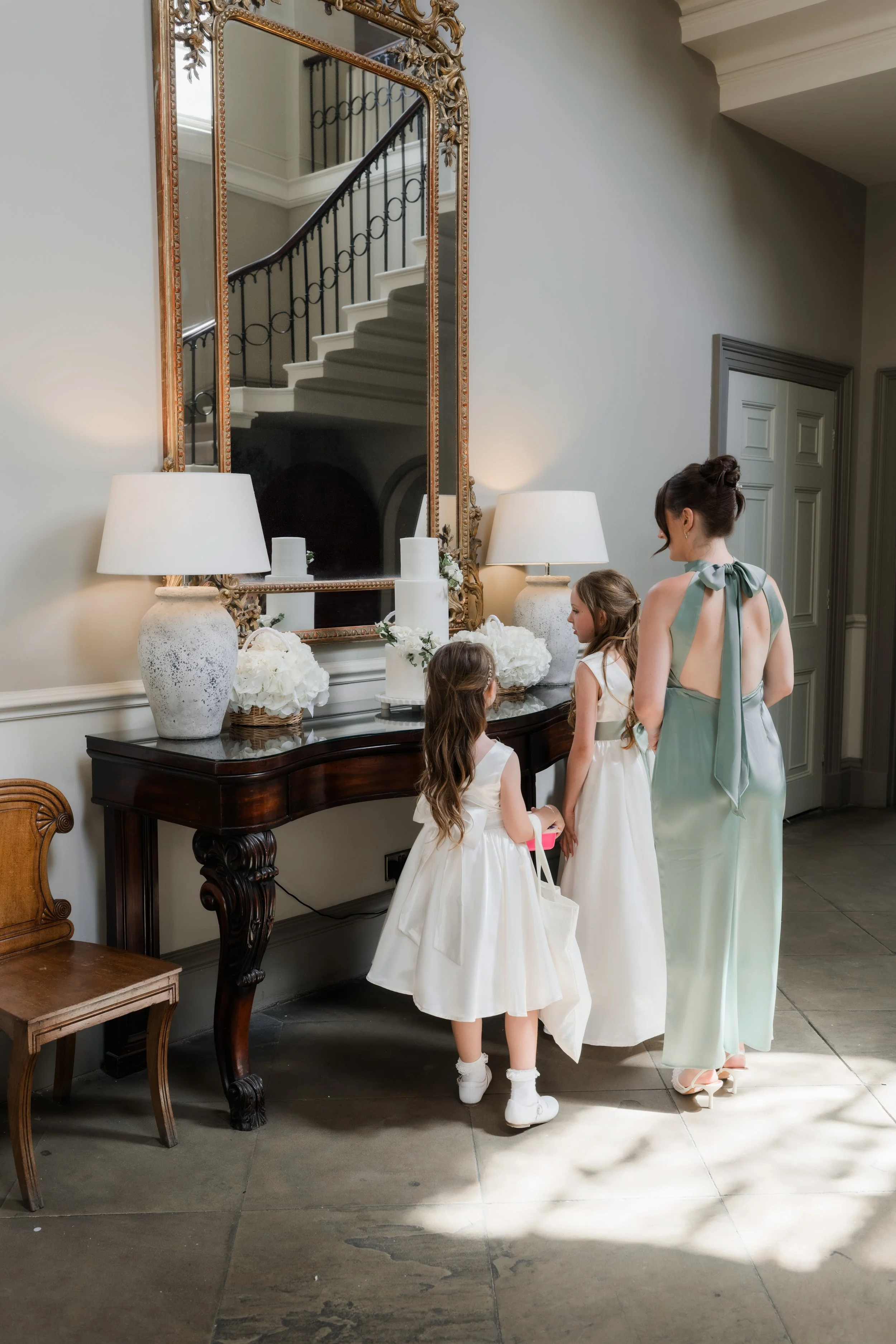 A woman in a light green dress and two young girls in white dresses are standing near a dark wooden table with three white lamps, white floral arrangements, and a decorative mirror on the wall. The mirror reflects a staircase with black wrought iron 