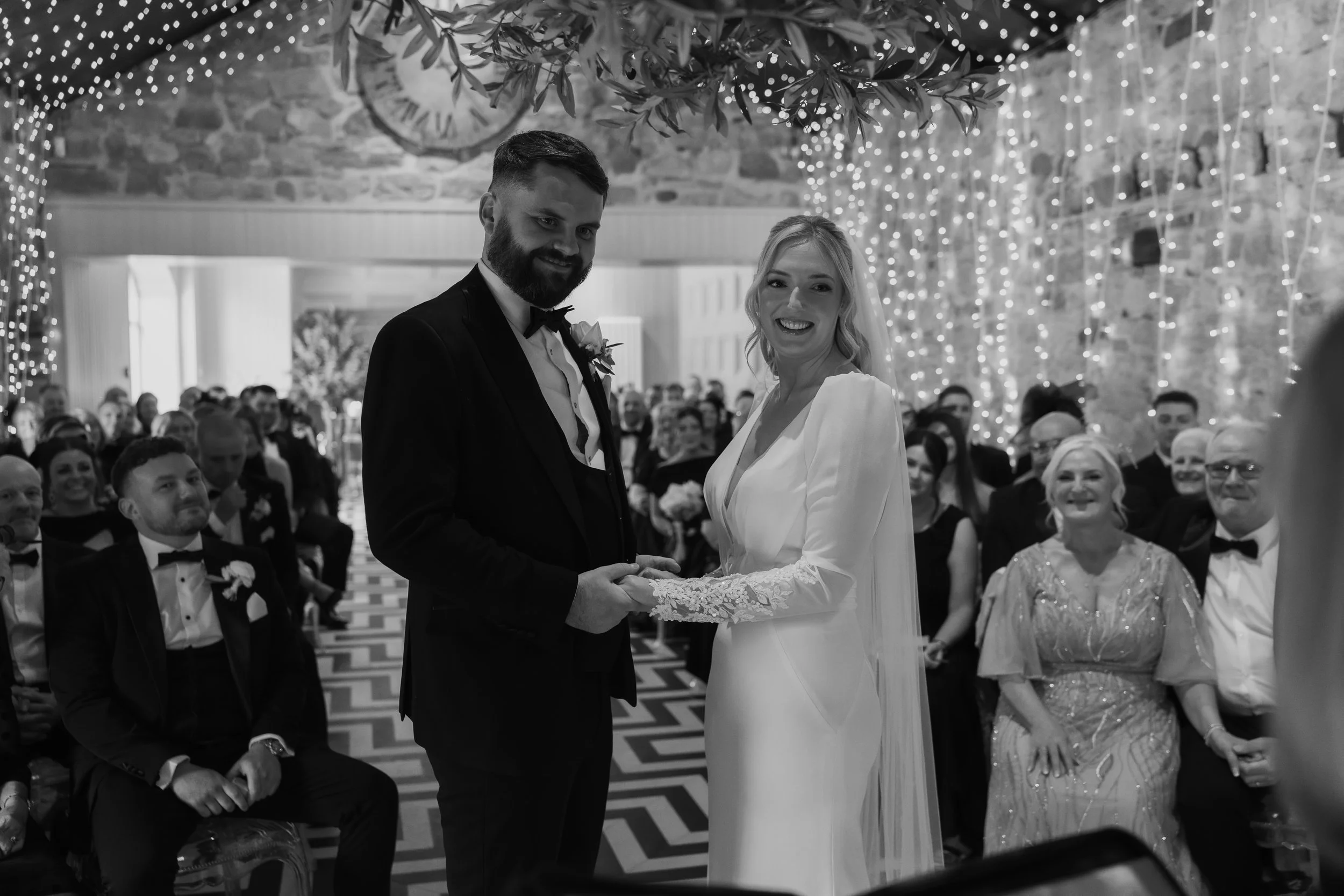 A black and white photo of a wedding ceremony. The bride and groom are holding hands and smiling at the camera. The ceremony is taking place indoors with guests sitting in the background, decorated with string lights and hanging foliage.