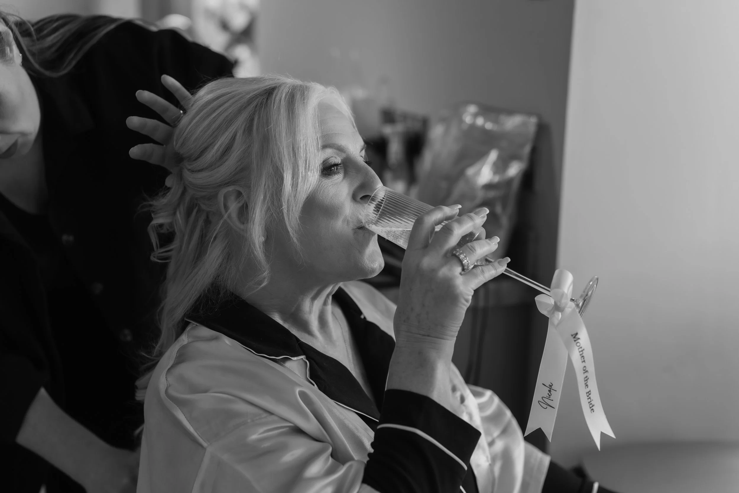 A woman with blonde hair sipping champagne from a flute, wearing a satin robe with a ribbon that reads "Mother of the Bride."