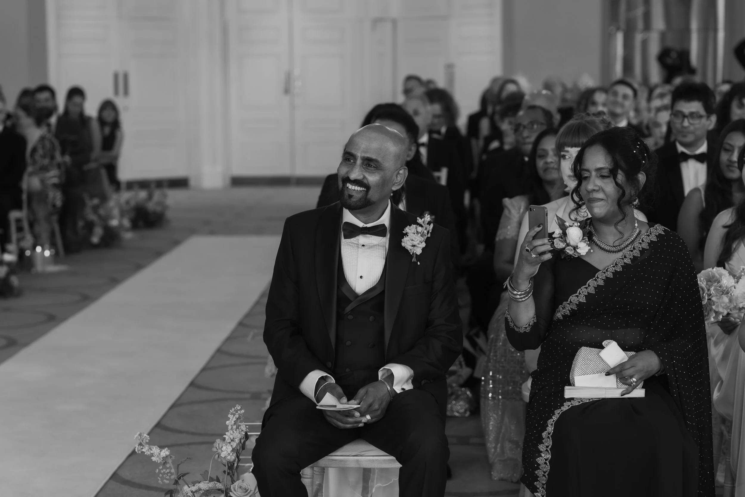 A black-and-white photograph of a wedding ceremony with a man in a tuxedo and a woman in traditional attire, sitting and watching the event, surrounded by guests.