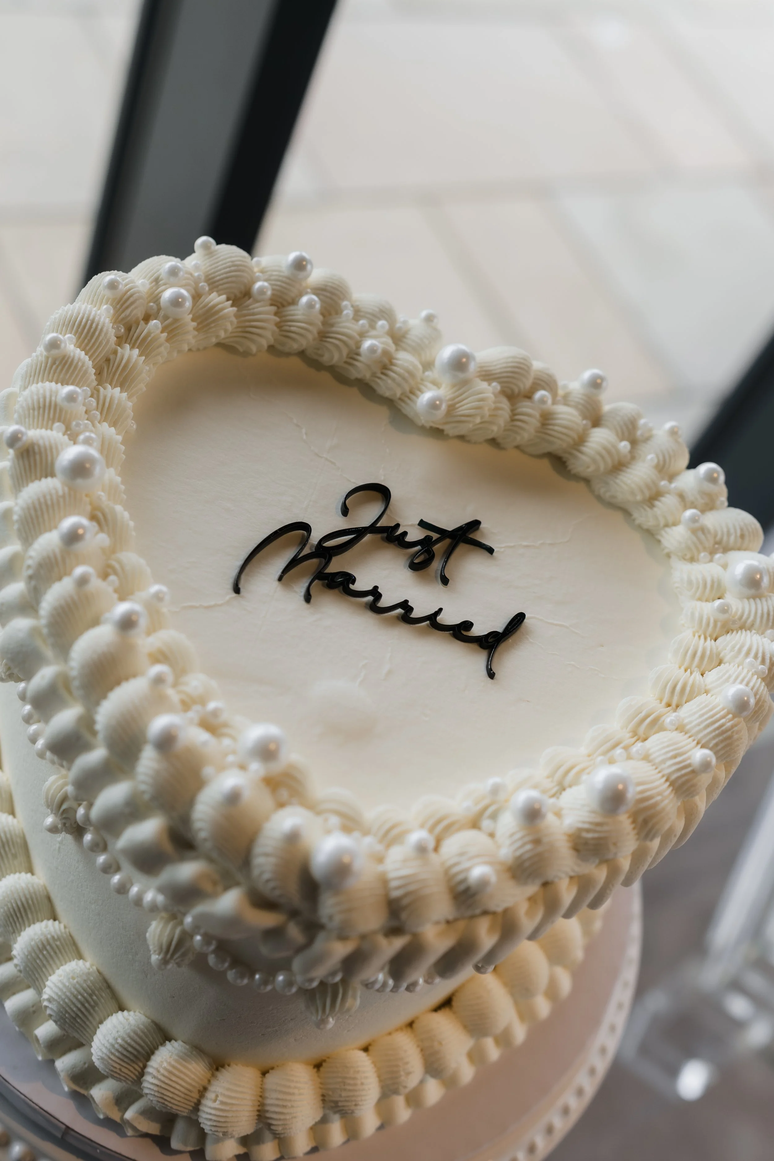 A white cake with a message that reads 'Just Married' in black letters, decorated with pearl-like beads and shell-shaped decorations around the edges.
