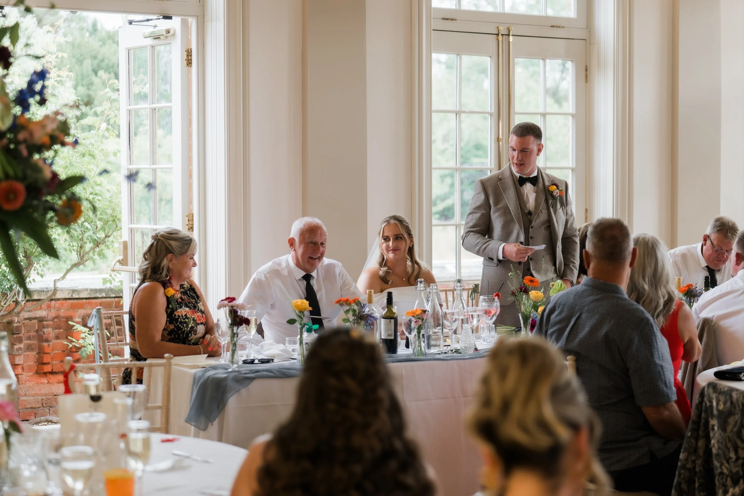 Groom giving a speech at a wedding reception, surrounded by seated guests and decorated with flowers.