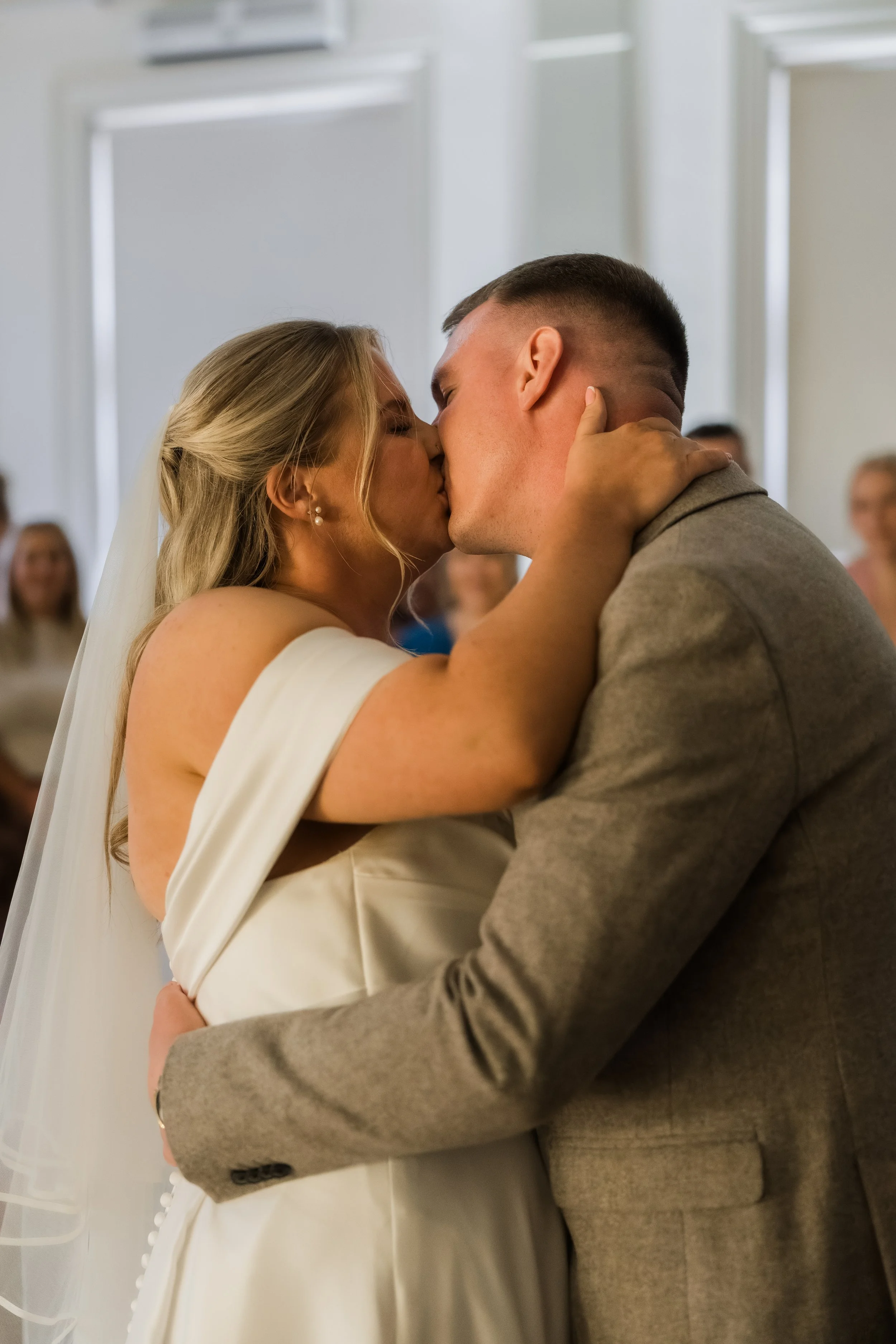 A bride and groom kissing during their wedding ceremony with guests watching in the background.