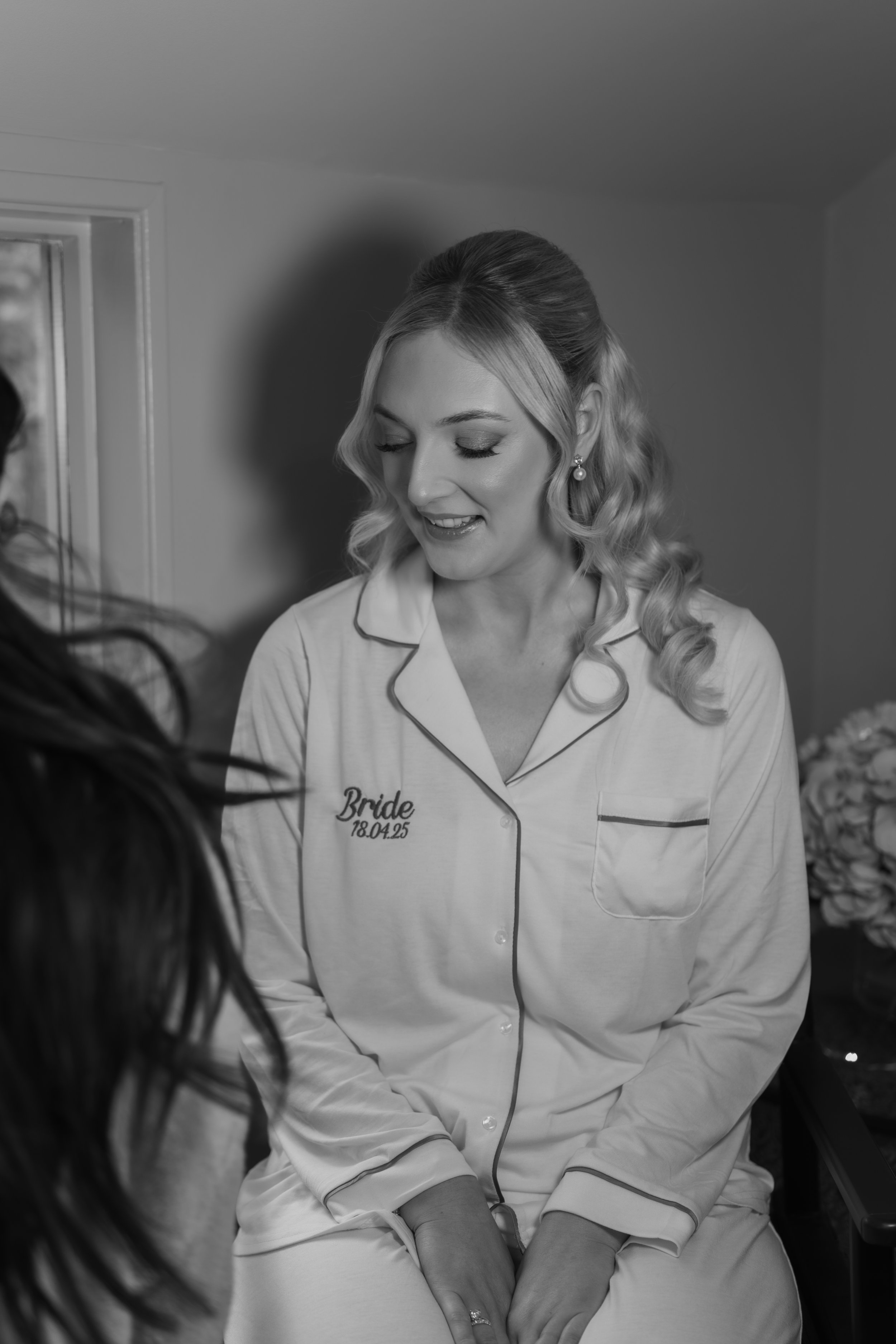 A bride with styled blonde hair and makeup wearing pajamas with "Bride" and date embroidered on it, smiling as she gets ready.