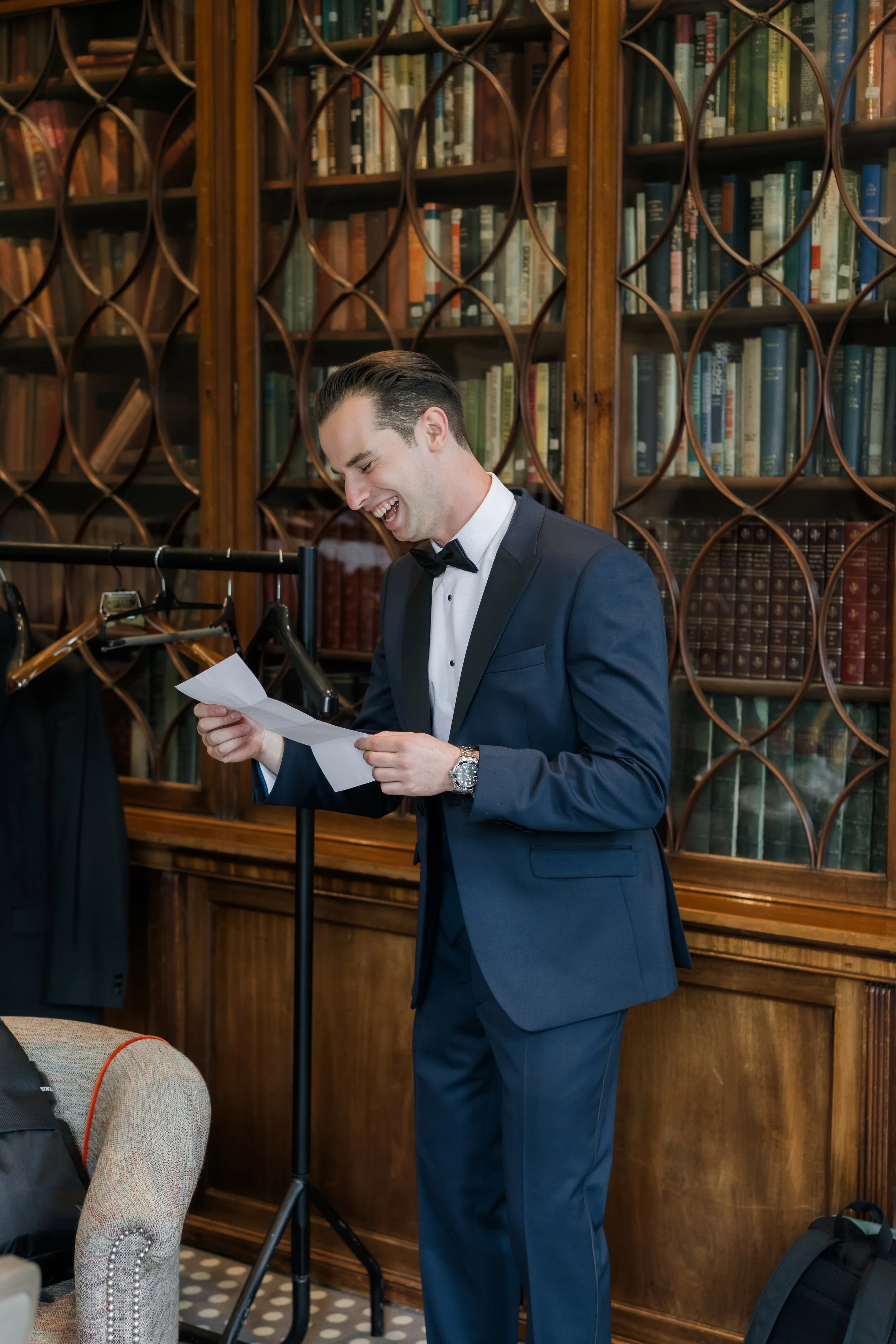 A man in a blue tuxedo with a black bow tie, smiling while reading a letter in a room with wooden bookshelves filled with books.