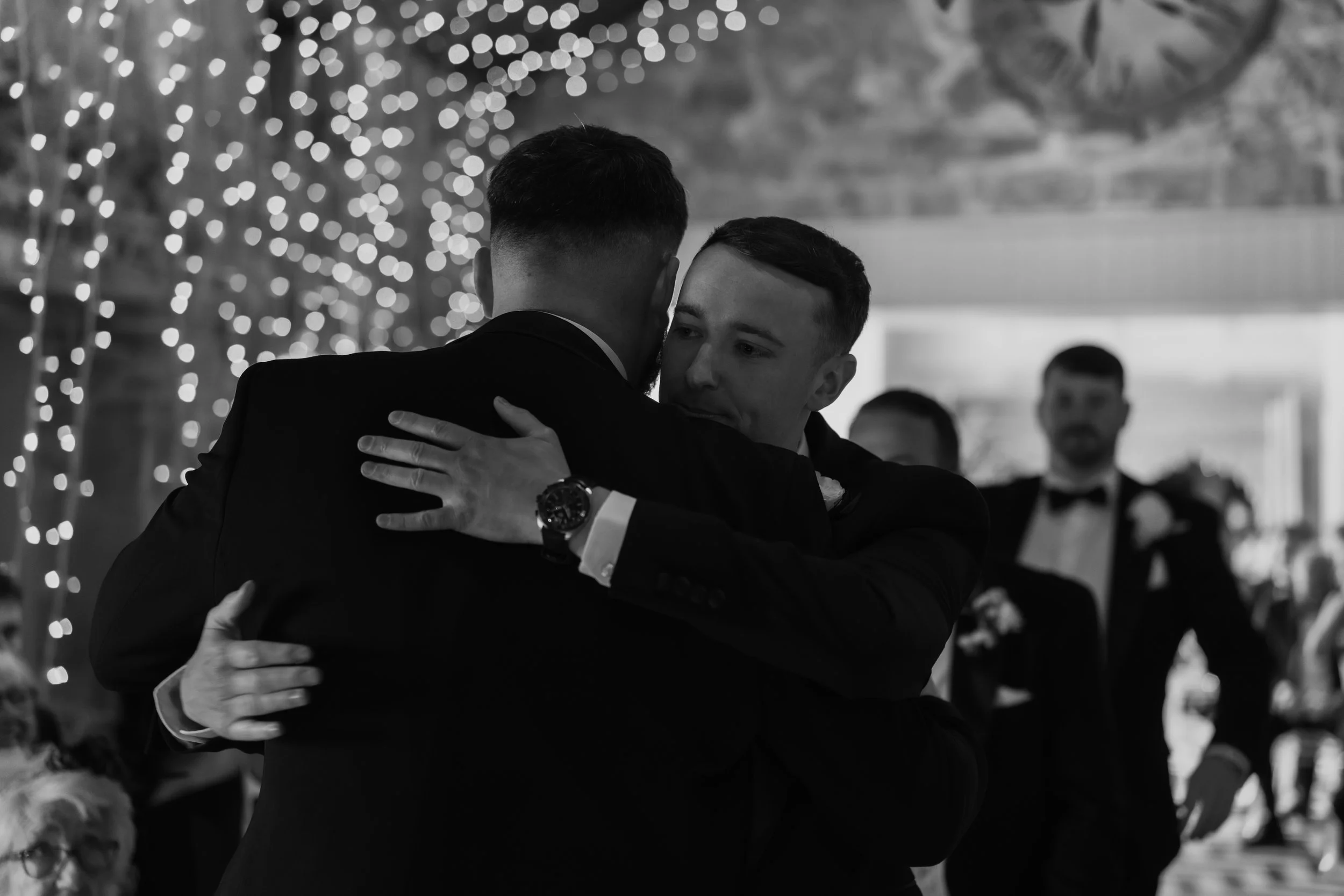 Two men in tuxedos share a hug at a wedding reception, with fairy lights and other guests in the background.