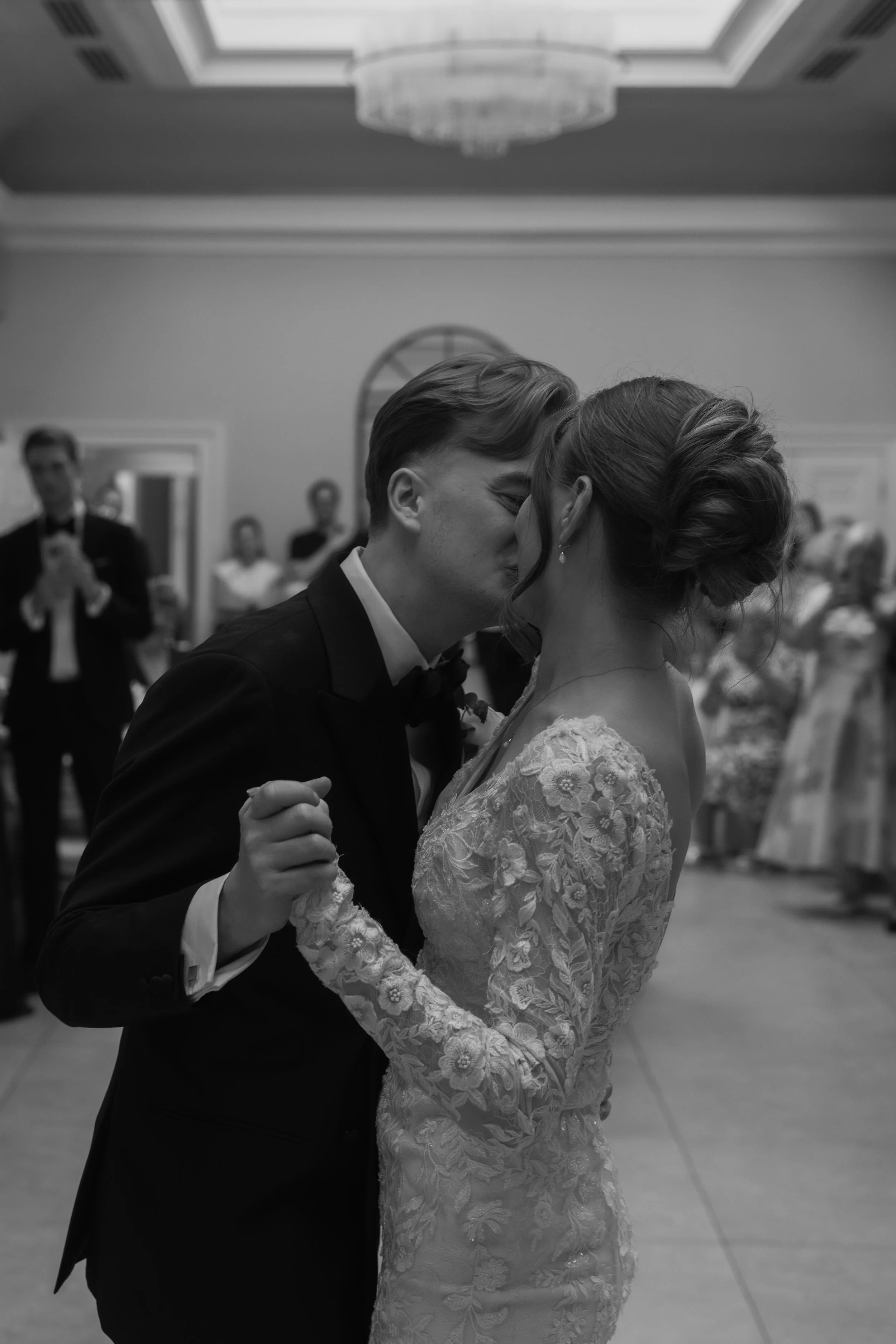 A black and white photo of a couple dancing closely at their wedding reception, with the groom in a tuxedo and the bride in a lace wedding gown, surrounded by guests in formal attire.