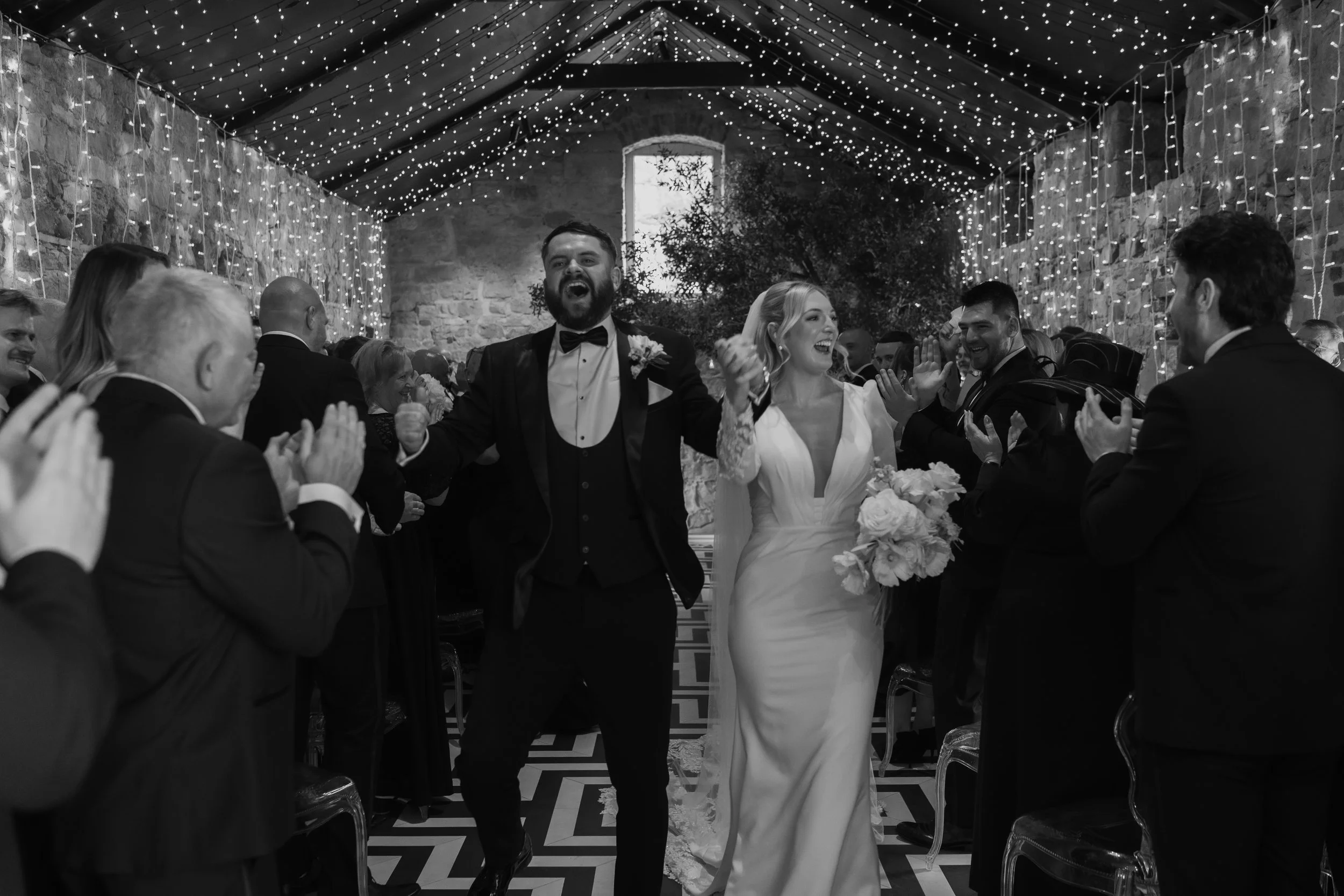 A newlywed couple walking down the aisle, hand in hand, smiling and celebrating in a wedding ceremony. The bride holds a bouquet of flowers, and the groom is dressed in a tuxedo. Guests are clapping and cheering on both sides. The setting is decorate