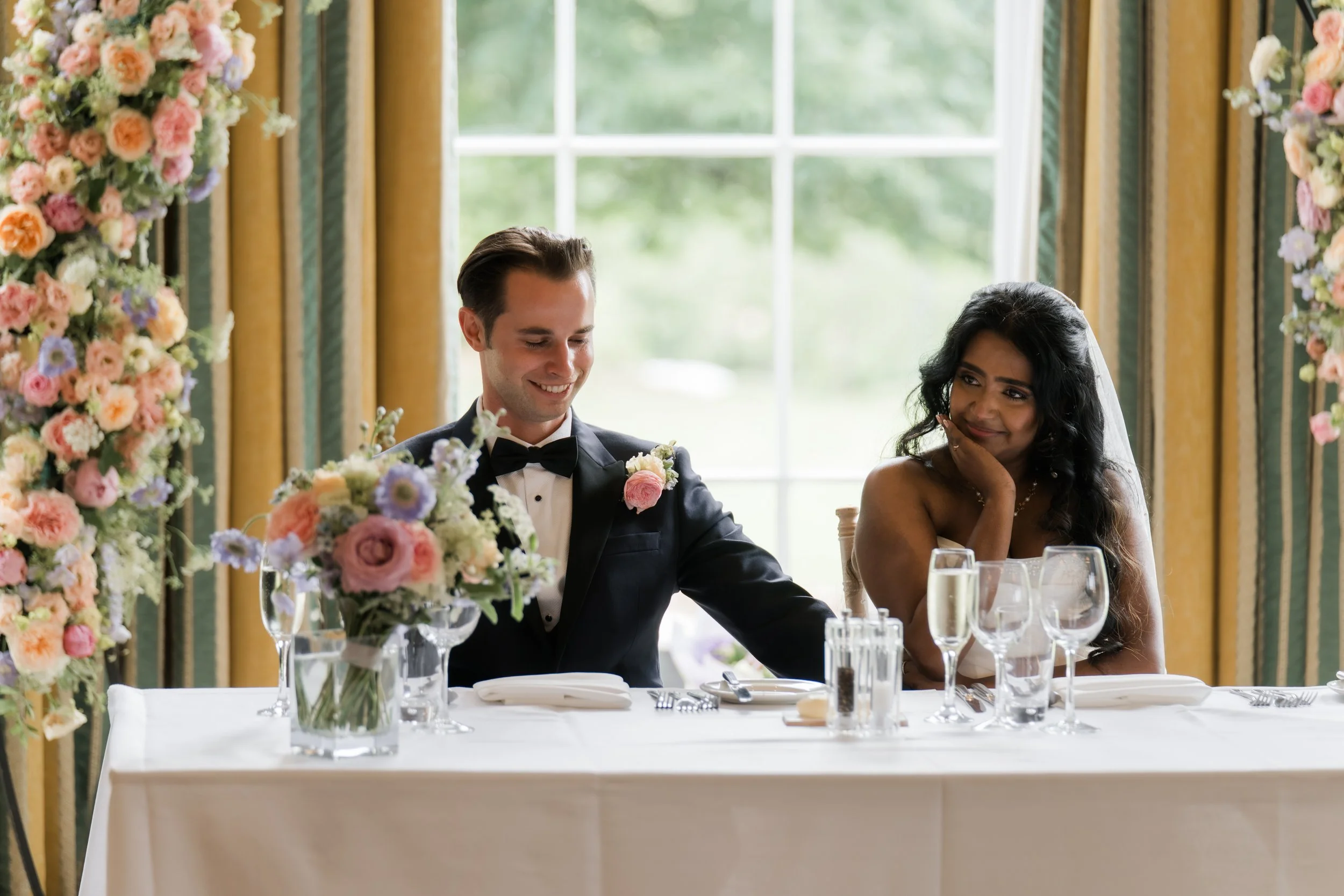 A bride and groom sitting at a wedding reception table with floral centerpieces, champagne glasses, and cutlery, smiling inside a decorated room with large window and yellow curtains.