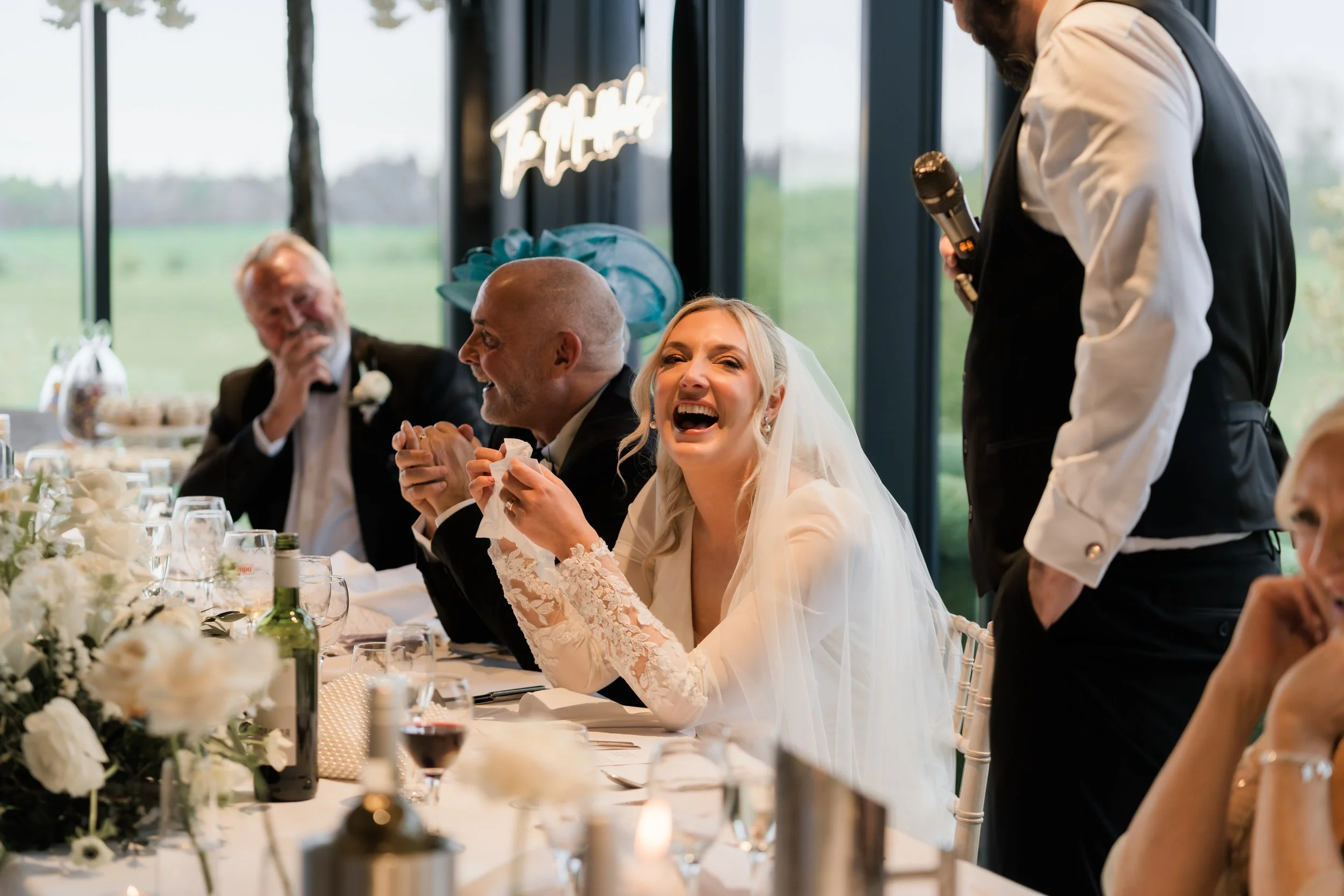 A bride laughing during a wedding reception, seated at a festive table with family, with a man standing and delivering speech holding a microphone.