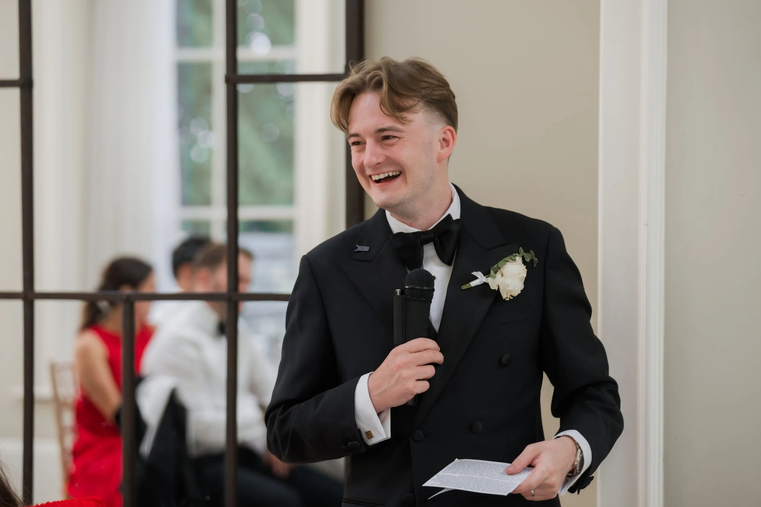 A smiling man in a tuxedo with a boutonniere holding a piece of paper, standing behind a black metal gate and speaking into a microphone, at what appears to be a formal event.