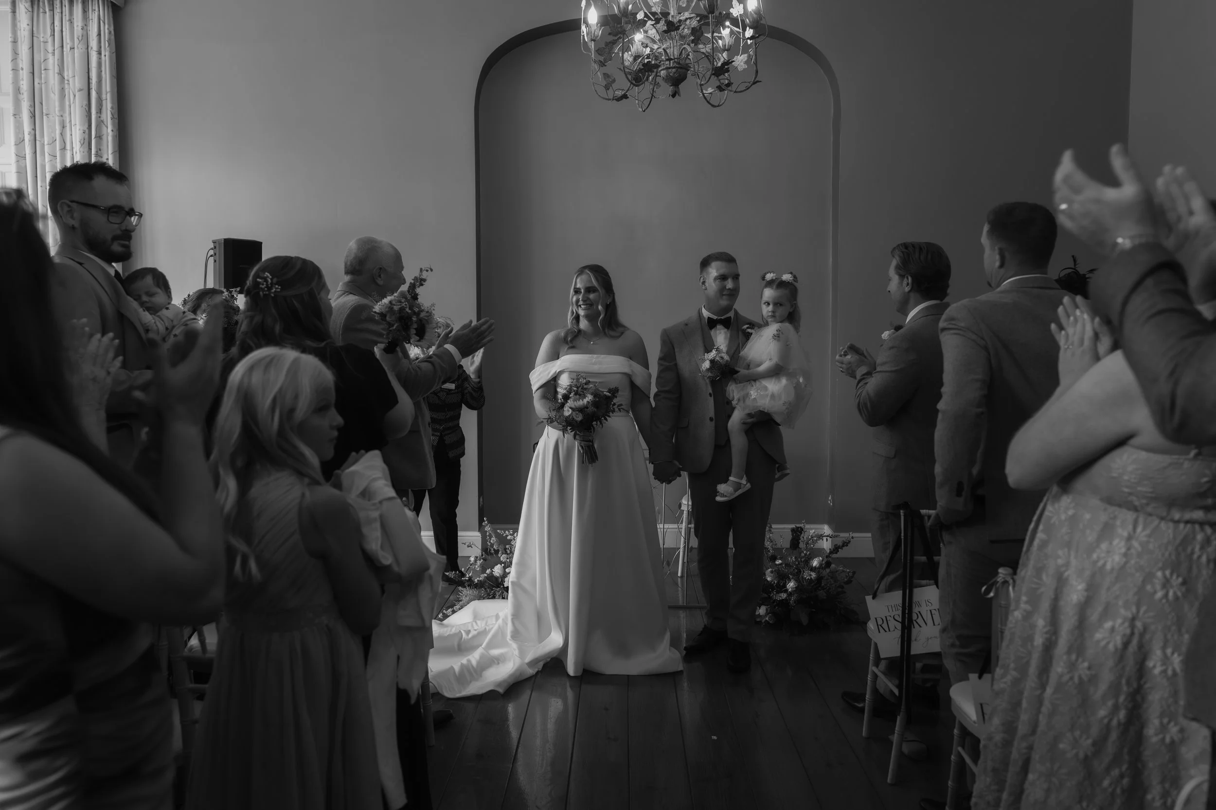 Black and white photo of a wedding ceremony with the bride and groom standing in the center, holding hands, surrounded by seated and standing guests clapping and celebrating.