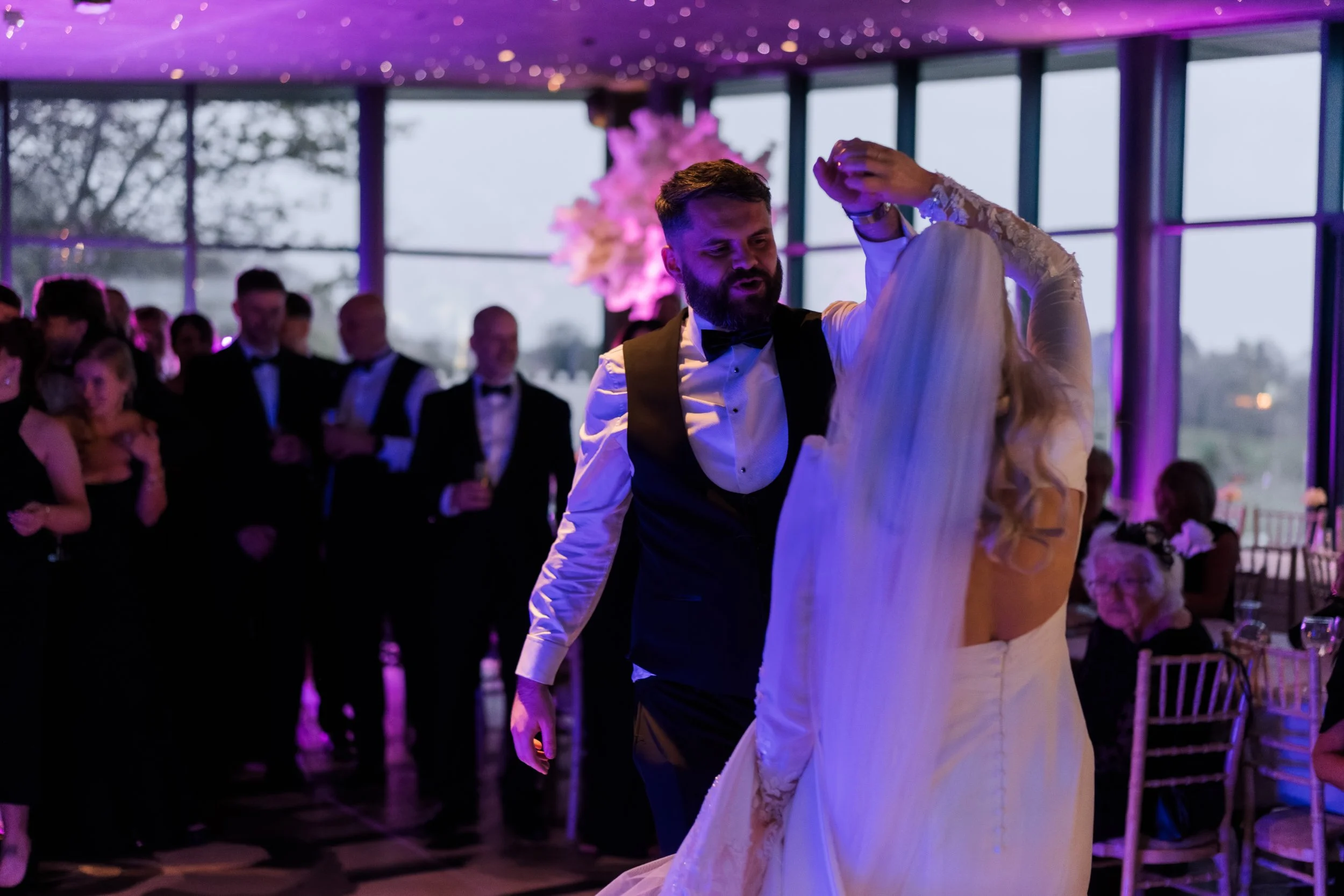 A man and woman dance at a wedding reception, with guests watching in the background, in a well-lit venue with large windows and purple lighting.