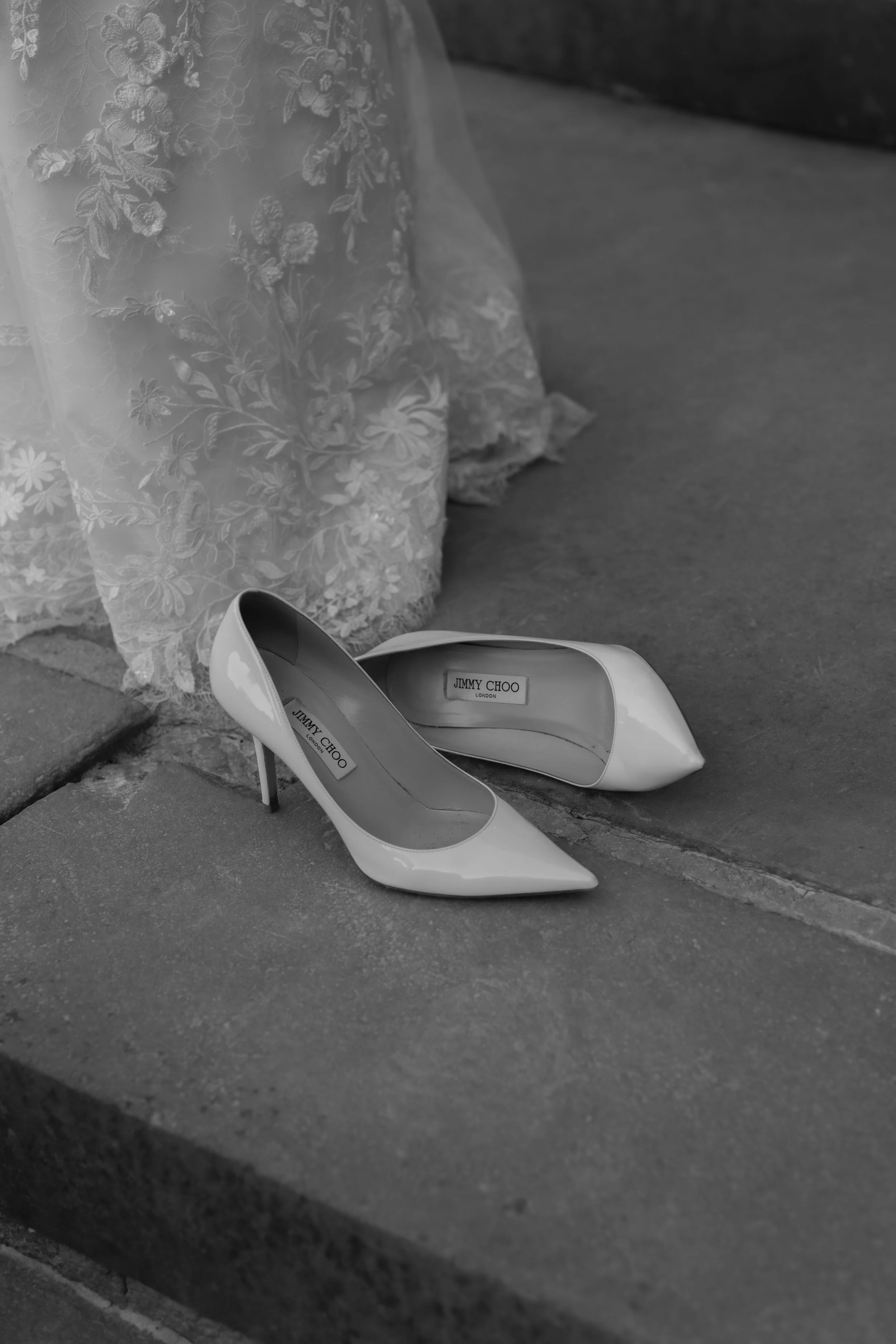 A pair of white Jimmy Choo high heels placed on a pavement in front of a wedding dress with floral embroidery.