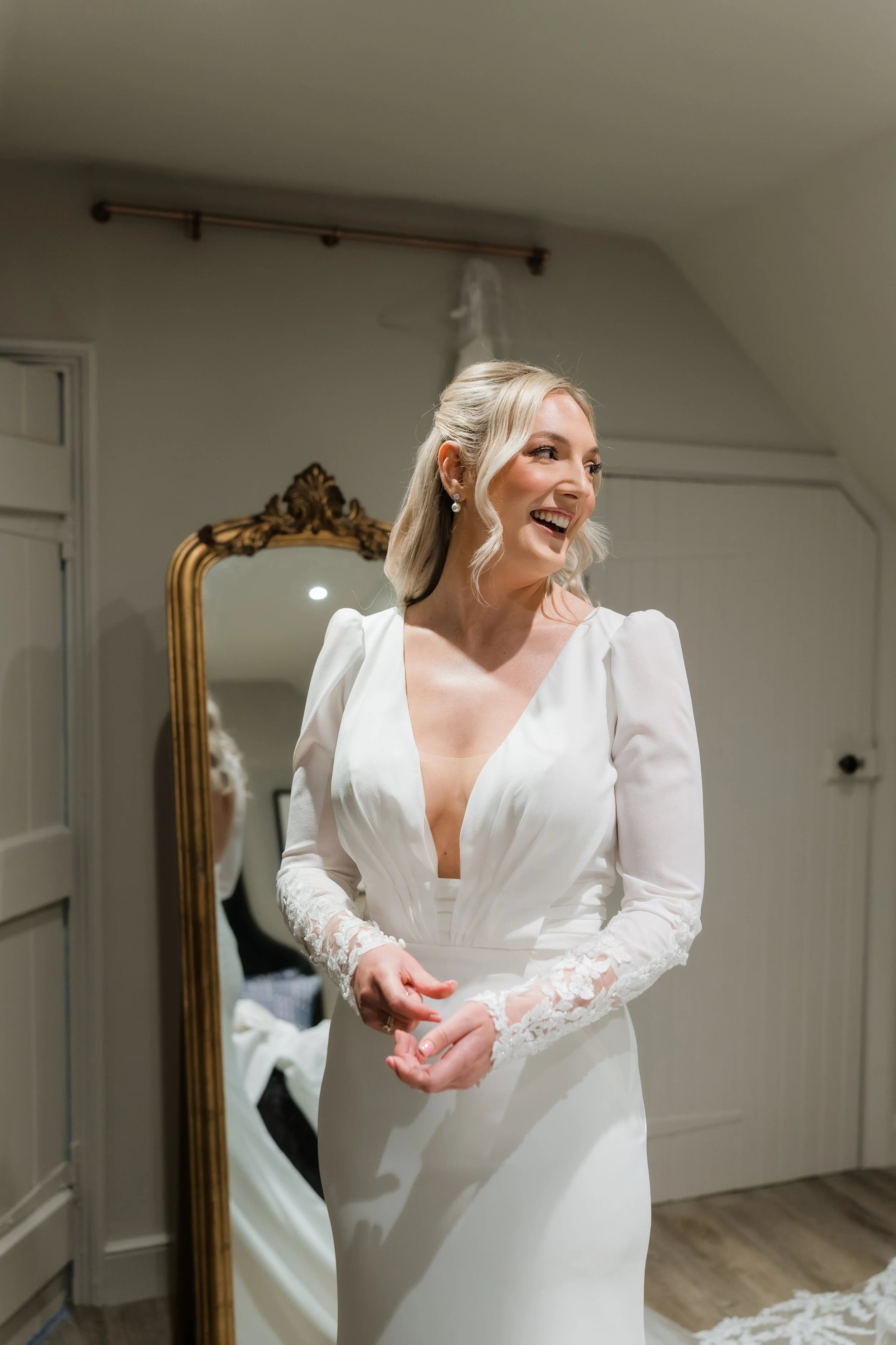 A woman in a white wedding dress smiling while looking to her right, standing in a room with a large mirror behind her.