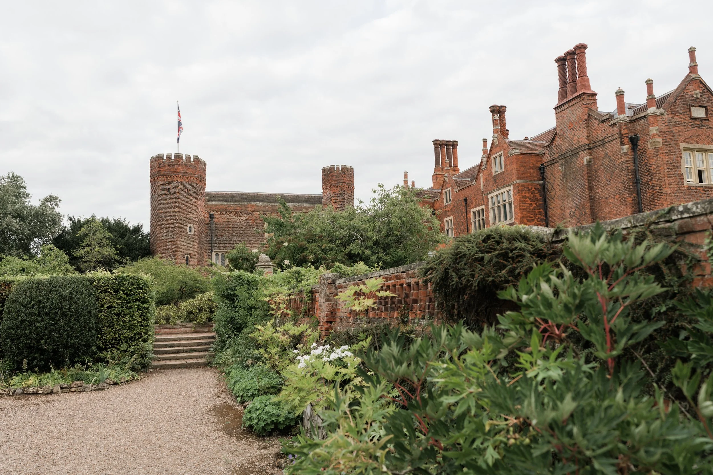 A historic red brick castle with multiple chimneys and a flag flying from a round tower. The castle is surrounded by lush greenery and a gravel pathway leading to stone steps.